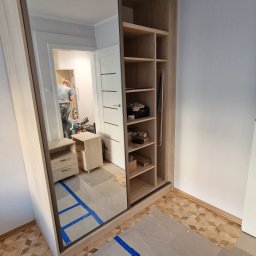 Fabryka mebli na wymiar - Montaż szafy wnękowej z lustrem i otwartymi półkami. Widoczne zabezpieczenia podłogi taśmą. W lustrze odbija się wnętrze pokoju z pracującym mężczyzną.