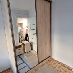 Fabryka mebli na wymiar - Jasna szafa przesuwna z lustrem w pokoju, widoczne zabezpieczenia podłogi. W lustrze odbija się wnętrze pokoju z osobami i meblami.