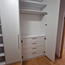 Fabryka mebli na wymiar - Biała szafa z czterema szufladami i drążkiem na ubrania, otwarte drzwi ukazują wnętrze. Minimalistyczny design, jasne oświetlenie, drewniana podłoga.