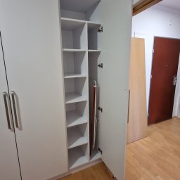Fabryka mebli na wymiar - Biała szafa z otwartymi drzwiami, widoczne półki i deska do prasowania w środku. Minimalistyczny design, jasne wnętrze, drewniana podłoga.
