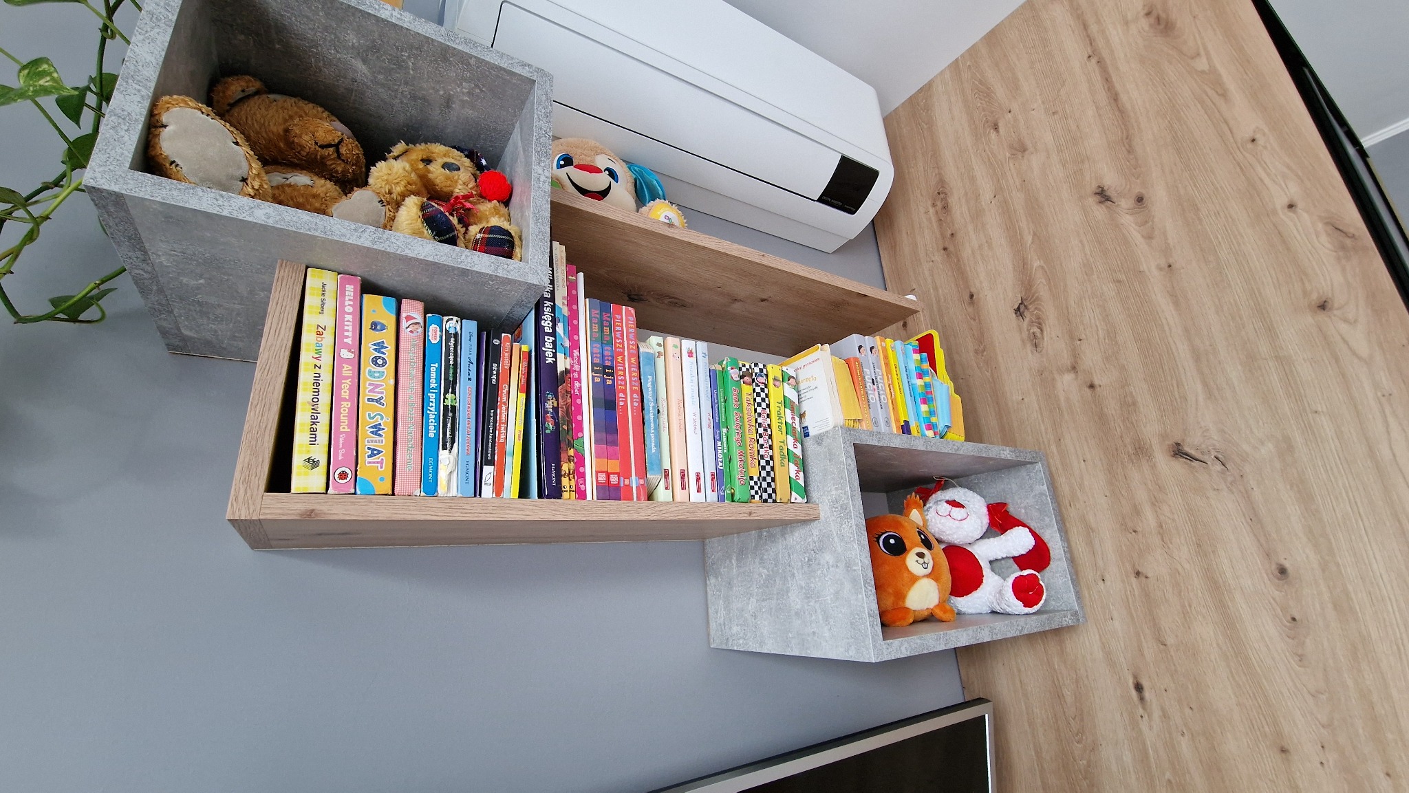 Nietypowa szafka na książki i zabawki dziecięce, z półkami w kształcie kostek w odcieniach szarości i drewna, zamontowana na szarej ścianie pod klimatyzatorem.