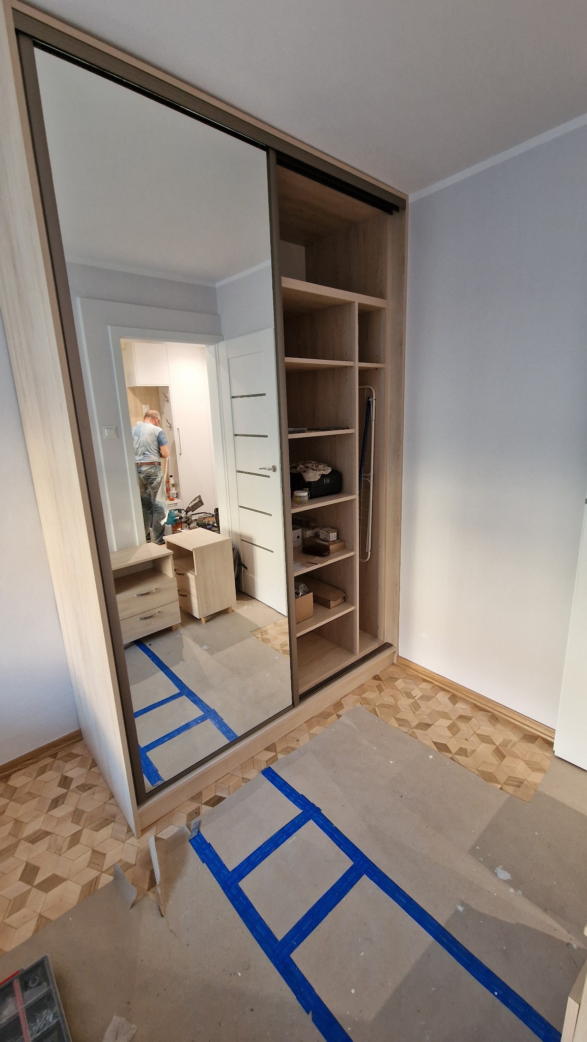 Montaż szafy wnękowej z lustrem i otwartymi półkami. Widoczne zabezpieczenia podłogi taśmą. W lustrze odbija się wnętrze pokoju z pracującym mężczyzną.