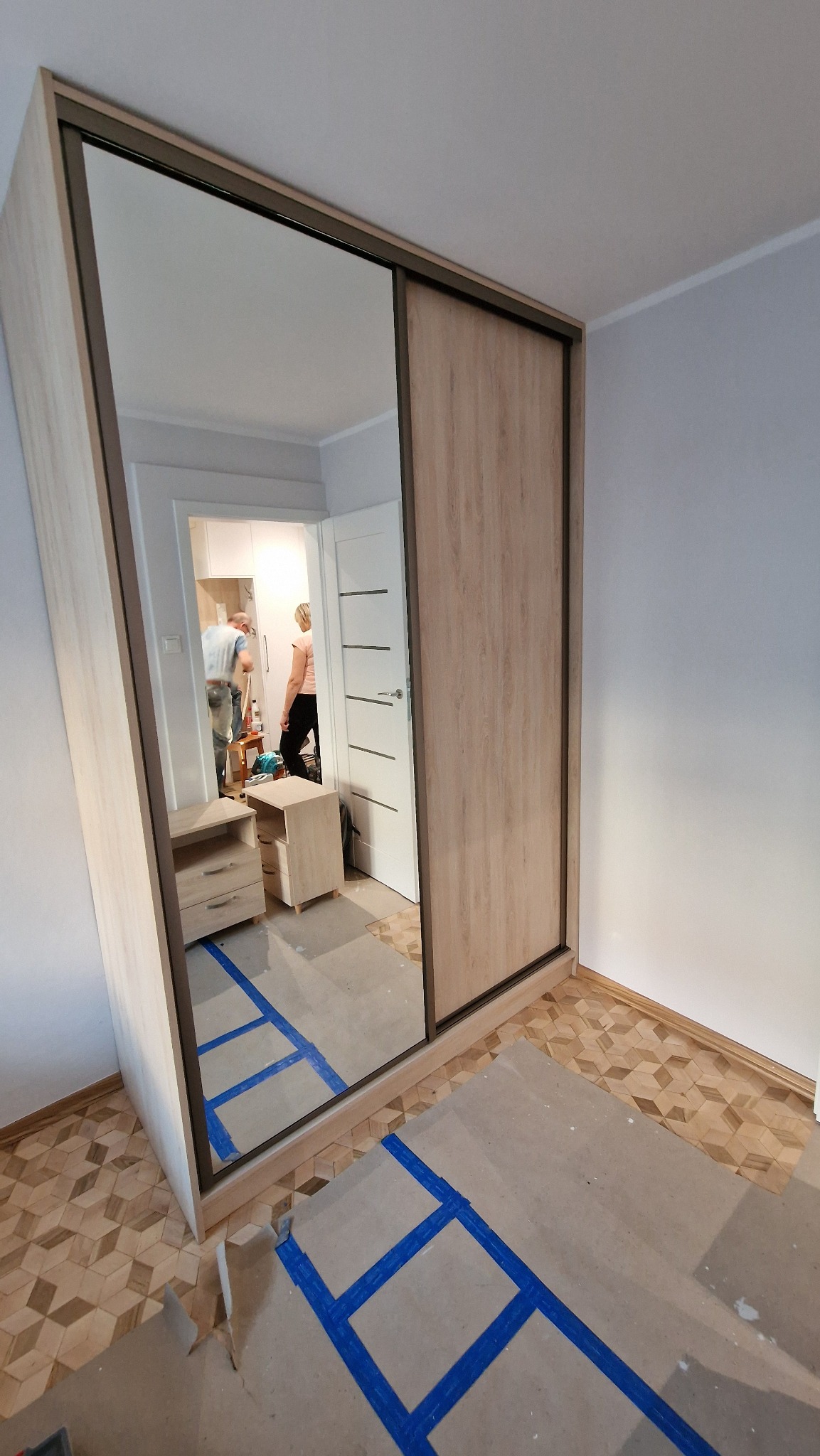 Jasna szafa przesuwna z lustrem w pokoju, widoczne zabezpieczenia podłogi. W lustrze odbija się wnętrze pokoju z osobami i meblami.