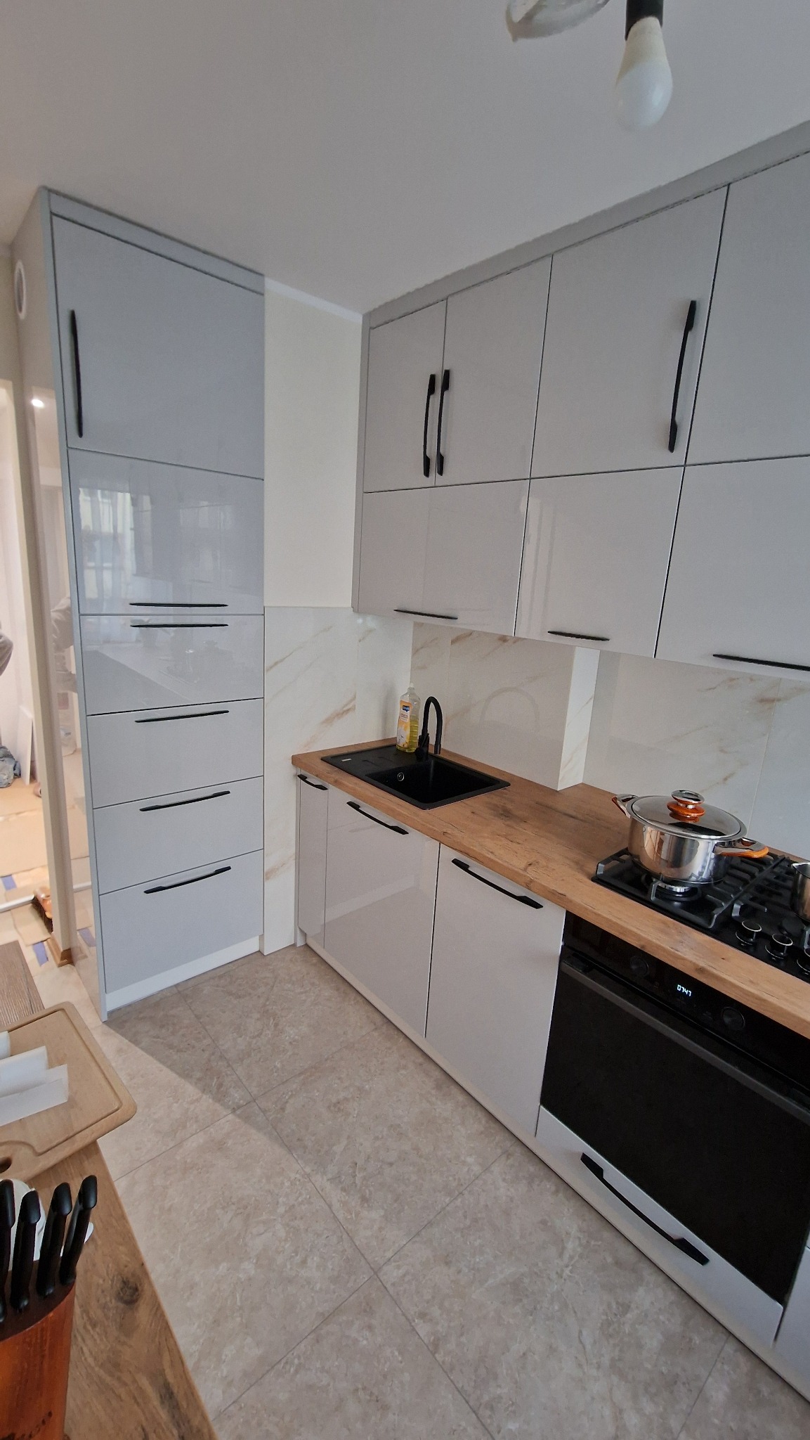 Nowoczesna kuchnia z szarymi, błyszczącymi frontami szafek i drewnianym blatem. Widoczny zlew, płyta grzewcza z garnkiem i wysoka szafka z szufladami.