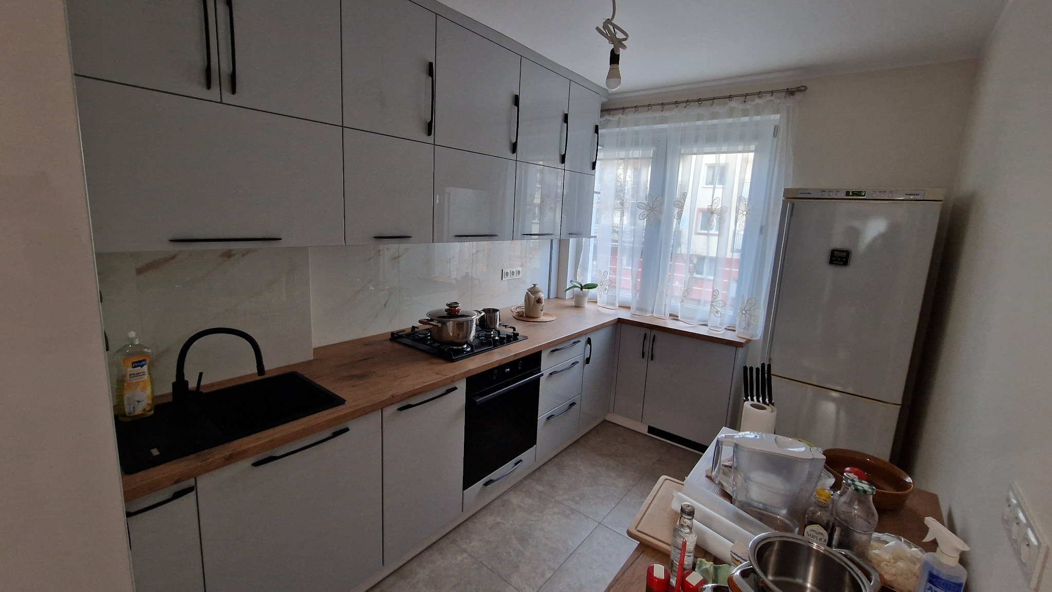 Jasnoszara kuchnia z drewnianym blatem, czarnym zlewem i płytą. Widoczne garnki na płycie, okno z firanką i lodówka. Ujęcie z perspektywy lekko z góry.