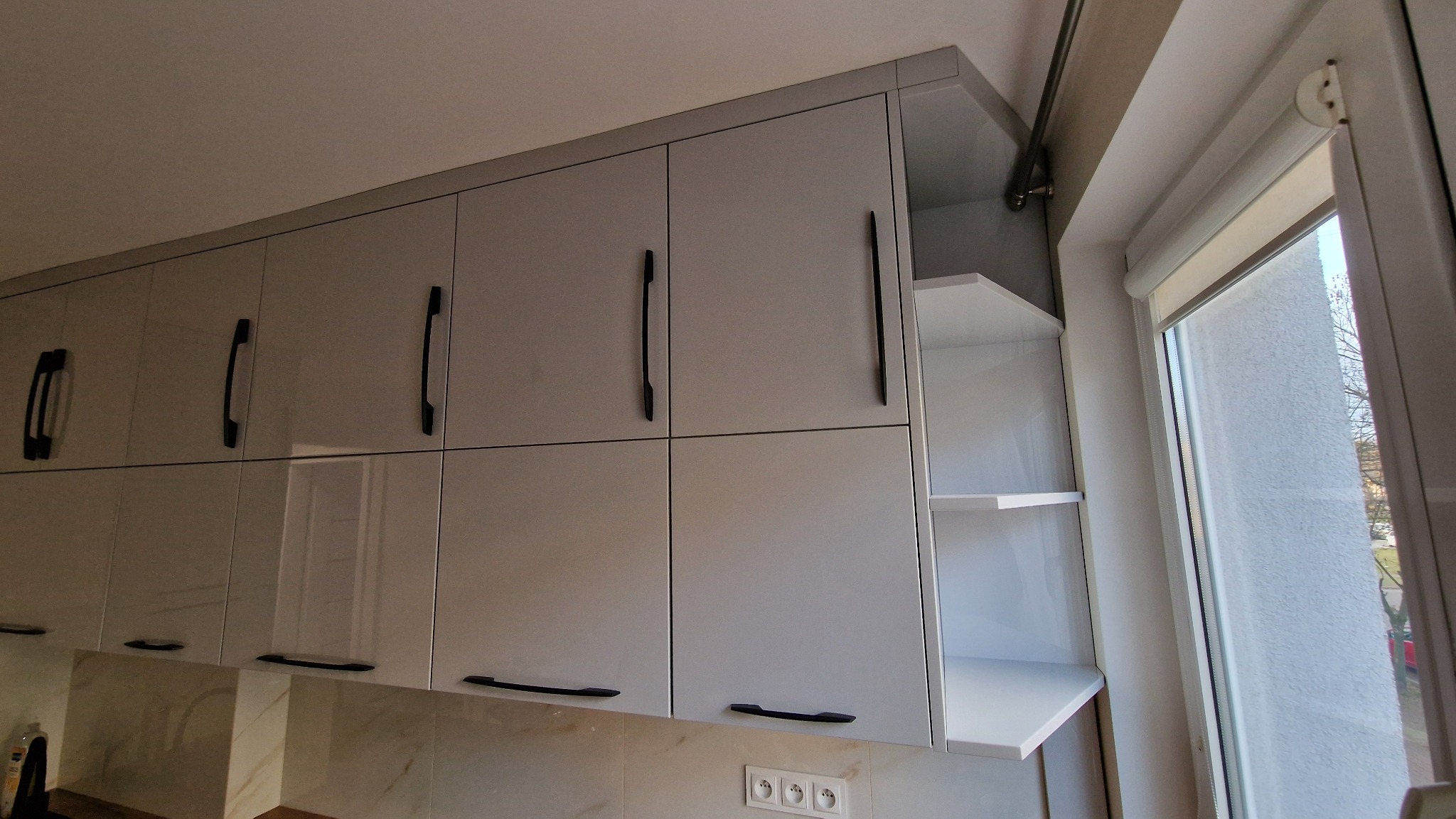 Jasnoszare szafki kuchenne z czarnymi uchwytami, część z otwartymi półkami, zamontowane pod sufitem, widok z dołu, obok okno z roletą.