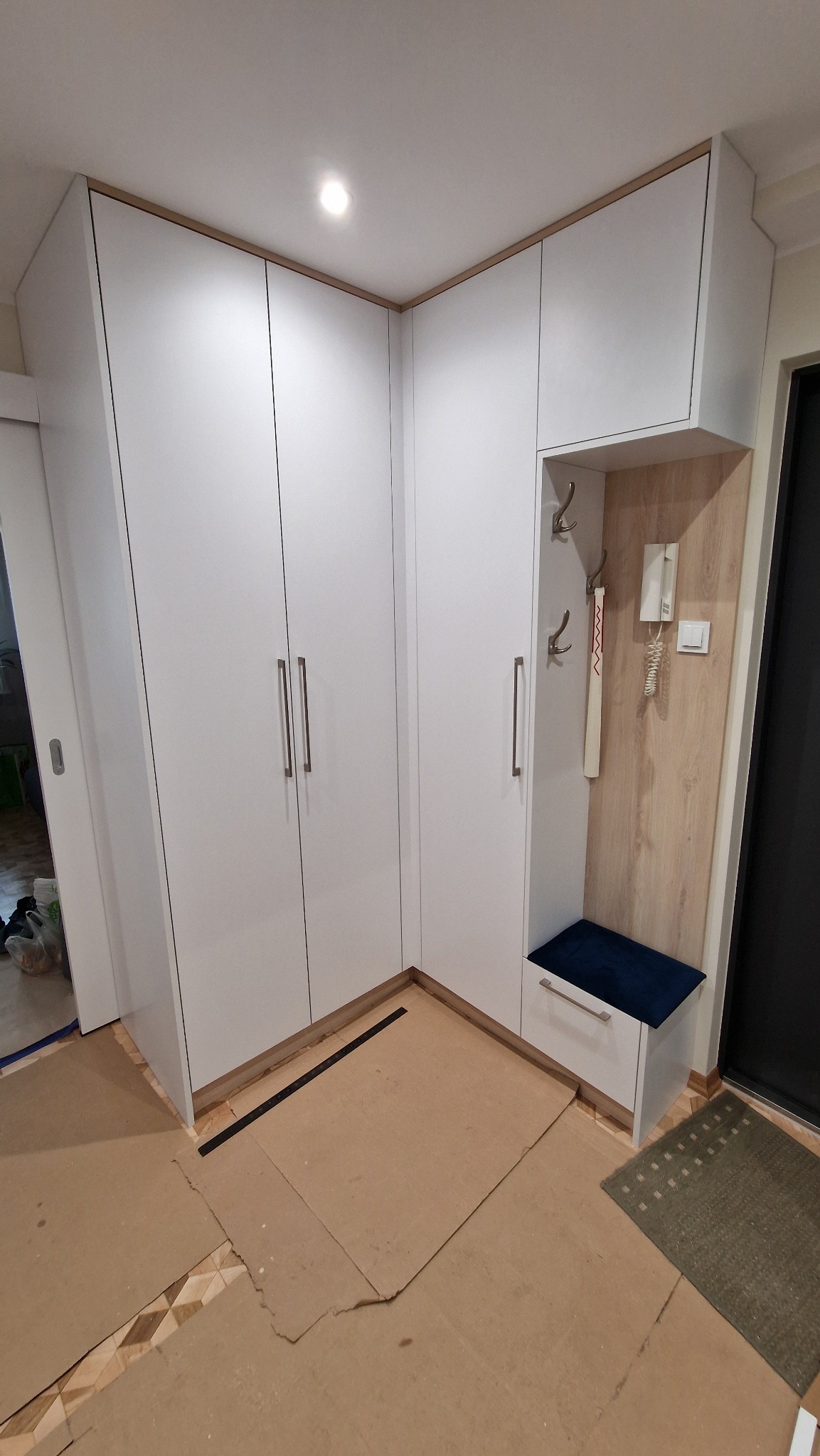 Biała, narożna szafa z siedziskiem i wieszakami w przedpokoju. Minimalistyczny design, jasne wnętrze, widoczne detale wykończenia. Na podłodze karton zabezpieczający.