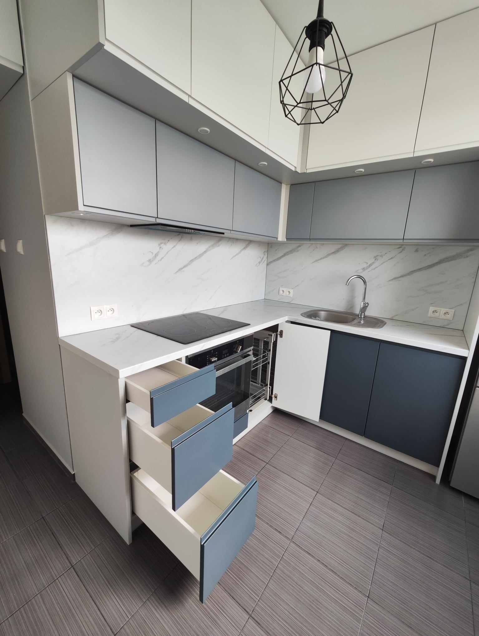 Nowoczesna kuchnia na wymiar z szarymi frontami, marmurowym blatem i otwartymi szufladami. Widoczny piekarnik i płyta indukcyjna. Minimalistyczna lampa sufitowa.