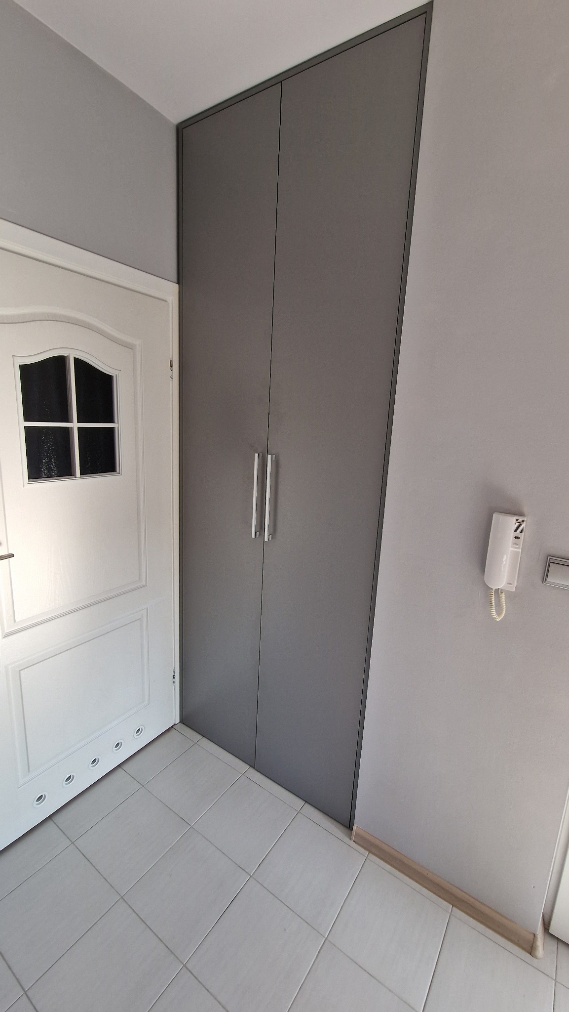 Szara szafa wnękowa z metalowymi uchwytami, dopasowana do koloru ścian, obok białych drzwi wejściowych i domofonu na tle jasnych płytek podłogowych.