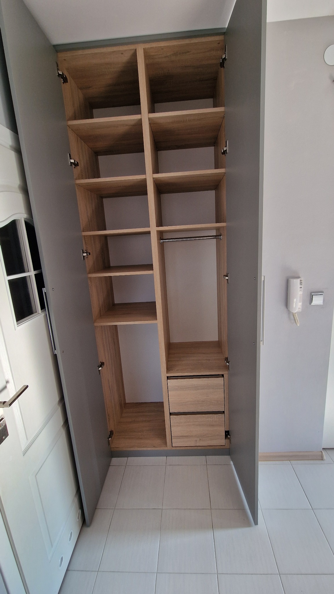 Otwarta szafa z półkami i szufladami w odcieniach drewna i szarości, widoczna z perspektywy wnętrza pomieszczenia, z drzwiami po bokach i białymi płytkami na podłodze.