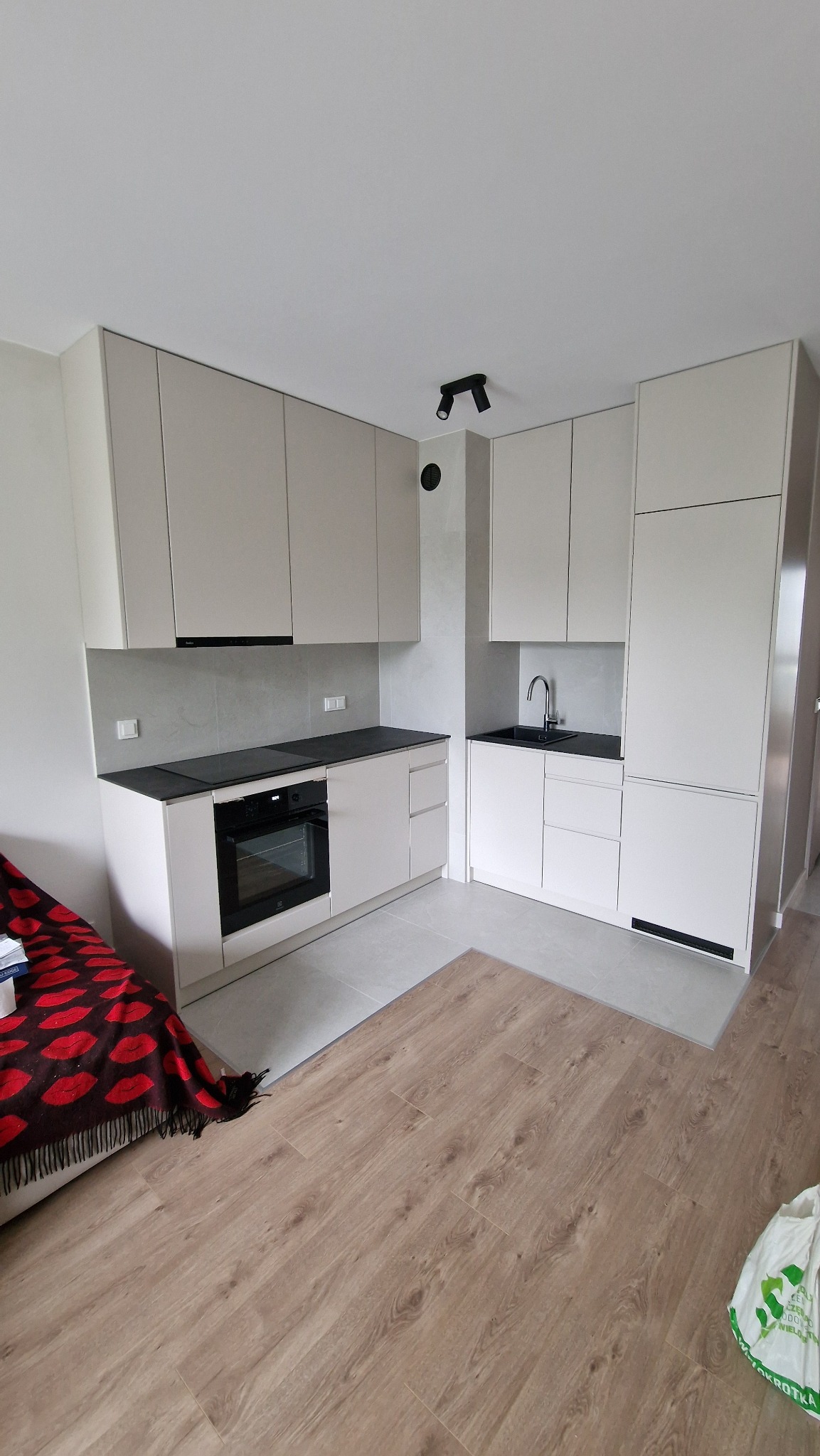 Jasna, nowoczesna kuchnia z zabudową pod sufit, czarnym blatem i płytą indukcyjną. Widoczne oświetlenie punktowe i fragment podłogi imitującej drewno.