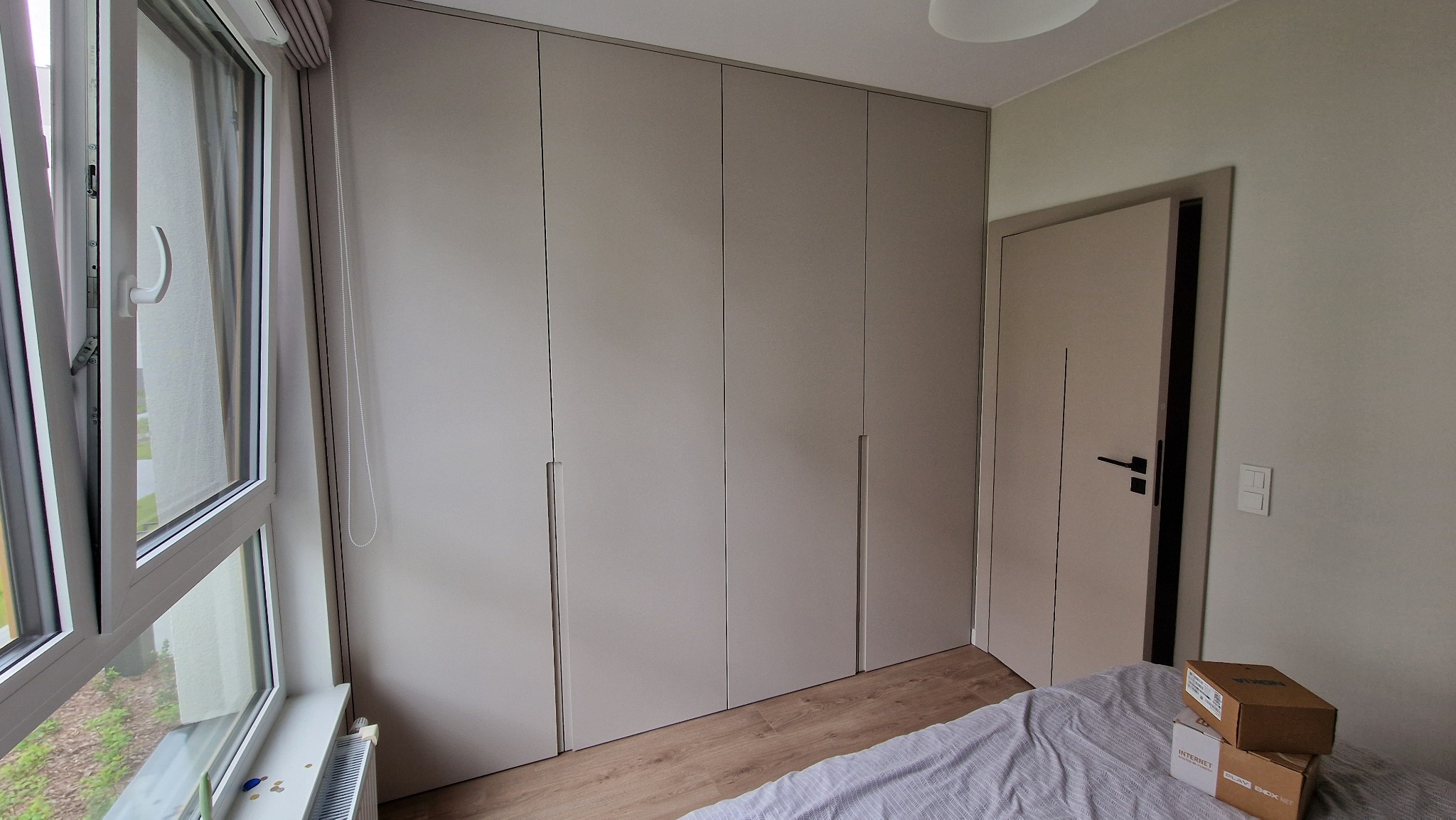 Beżowa szafa wnękowa z minimalistycznymi uchwytami, dopasowana kolorystycznie do drzwi i ścian. Obok okno z roletą i fragment łóżka z kartonami.