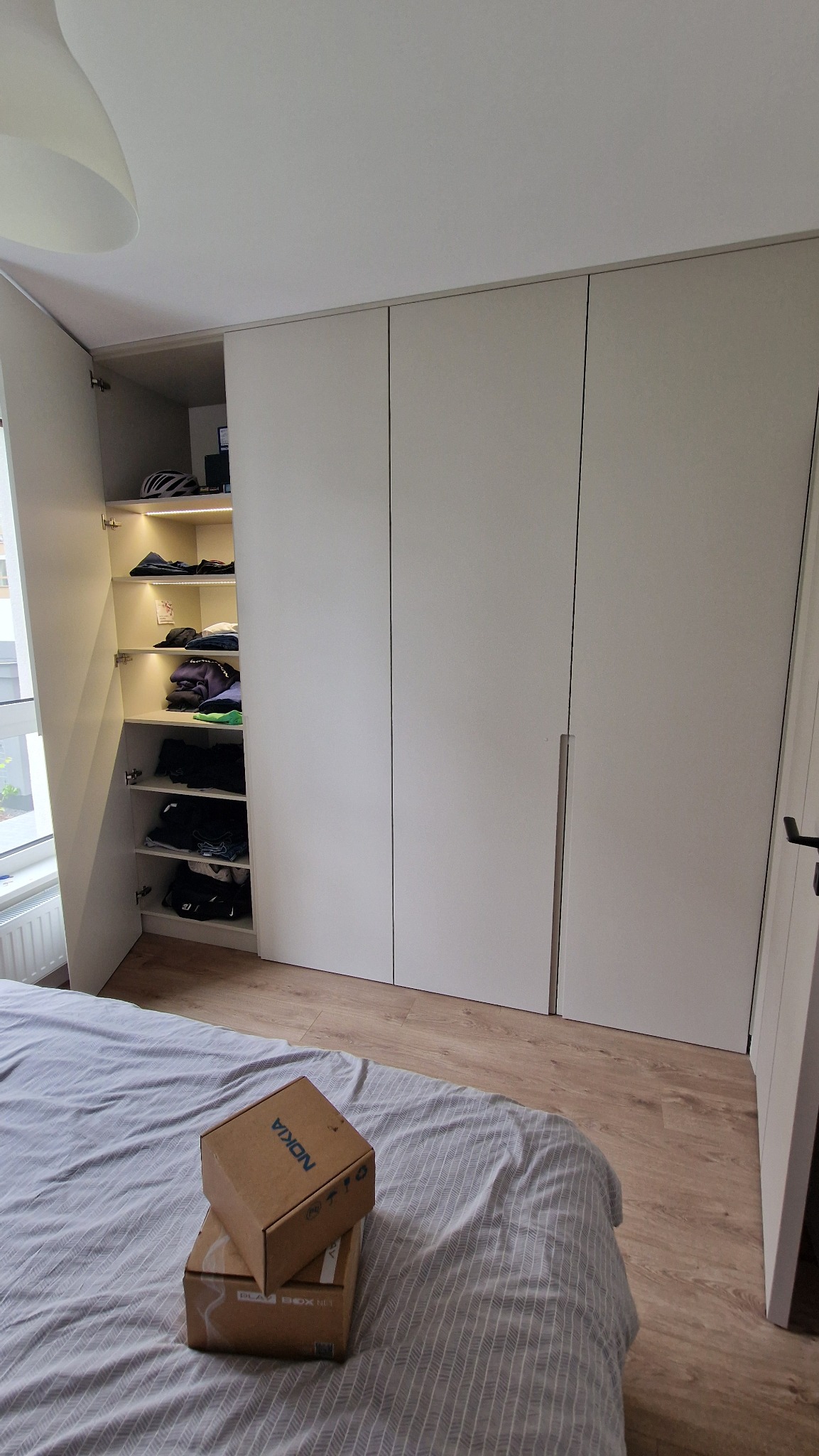 Biała szafa wnękowa z oświetleniem LED wewnątrz, otwarte drzwi ukazują półki z ubraniami. Na łóżku leżą dwa kartony.