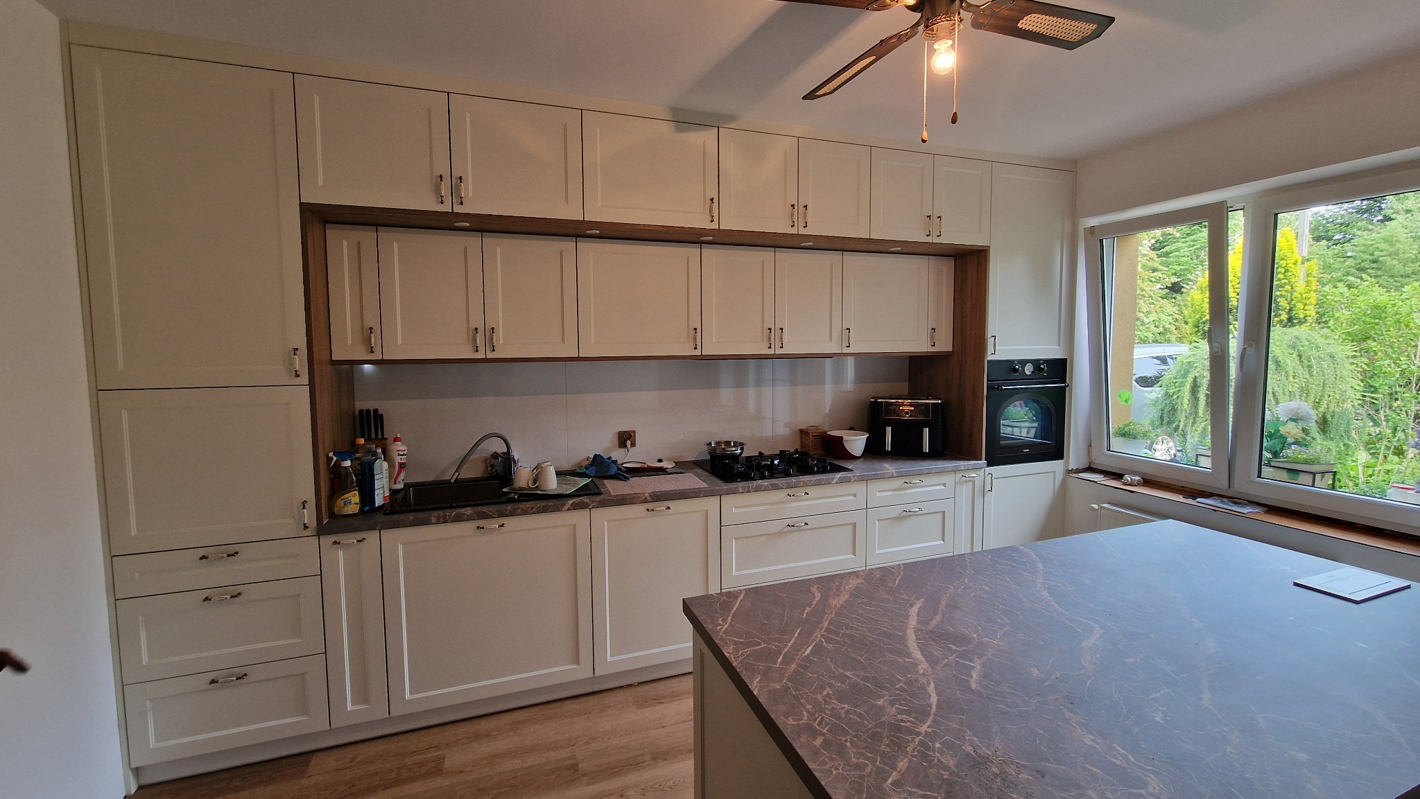 Jasna kuchnia na wymiar z wyspą i drewnianymi akcentami. Widok na ogród przez duże okno. Nowoczesne wykończenie, marmurowy blat wyspy.