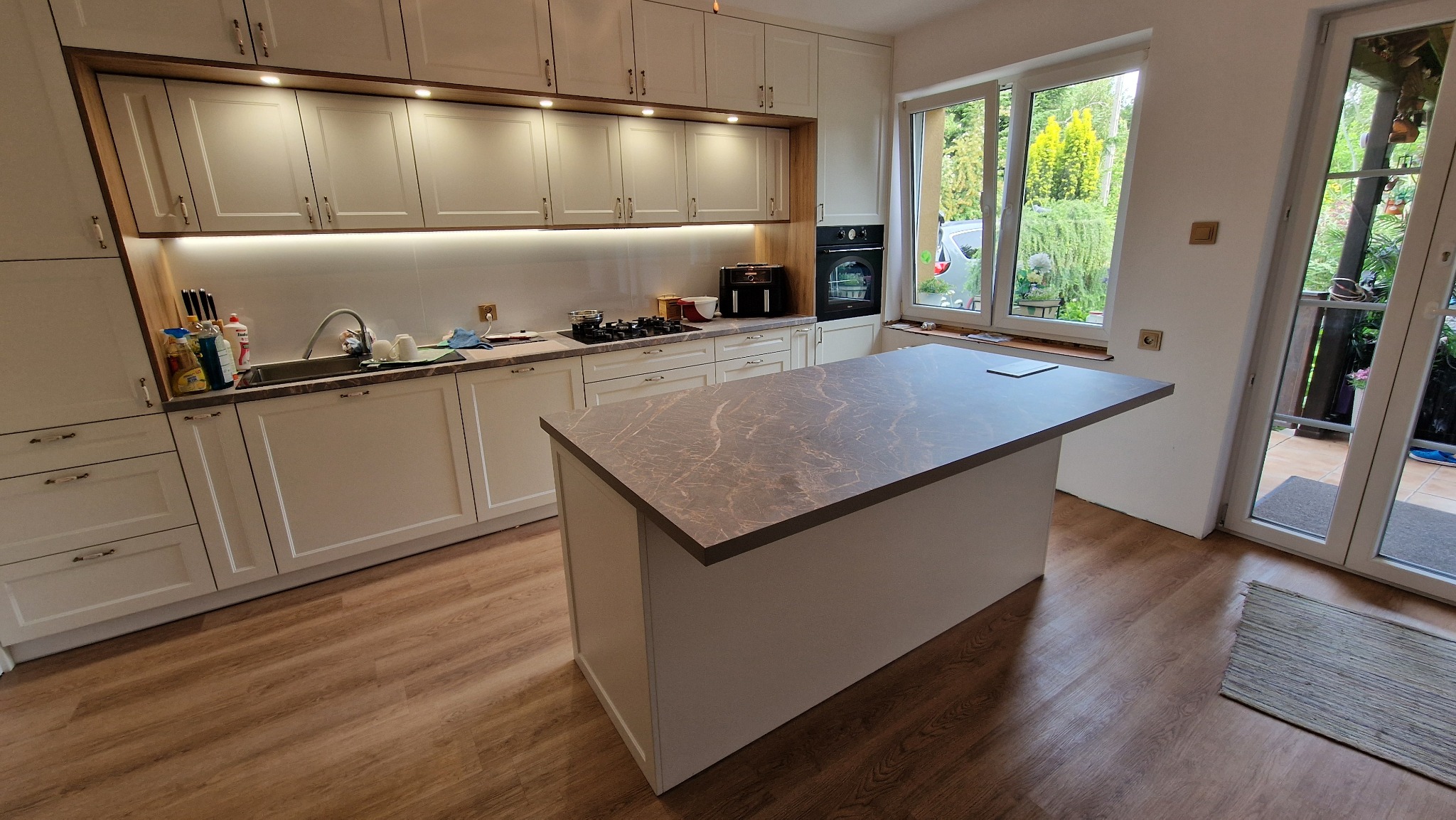 Jasna kuchnia z wyspą i blatem w odcieniach szarości. Widoczne szafki górne z oświetleniem LED, okno z widokiem na zieleń i drewniana podłoga.