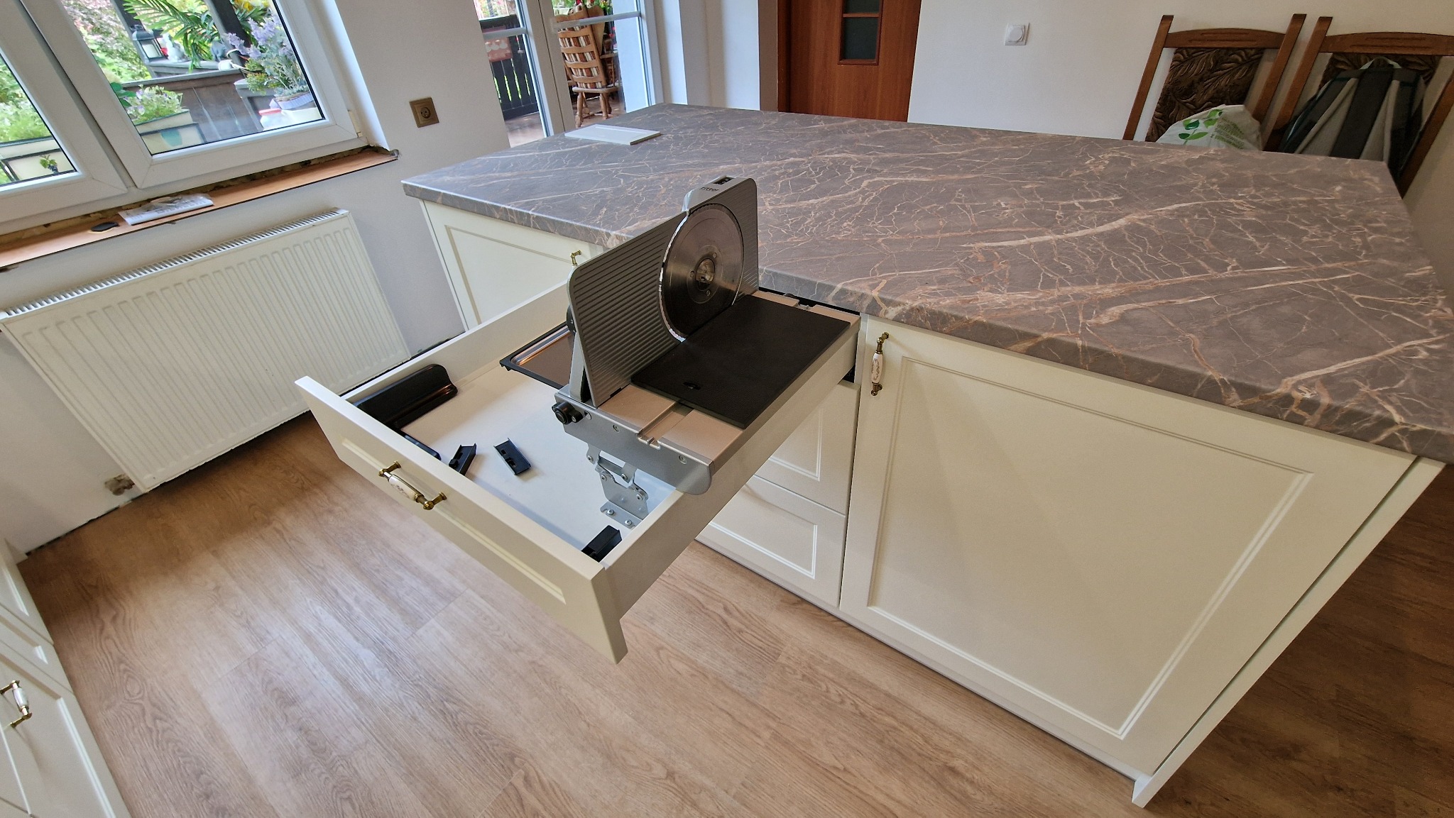 Kuchenna wyspa z blatem imitującym marmur i wysuwaną szufladą, w której zamontowano krajalnicę do wędlin. Widok z góry na jasne fronty szafek.