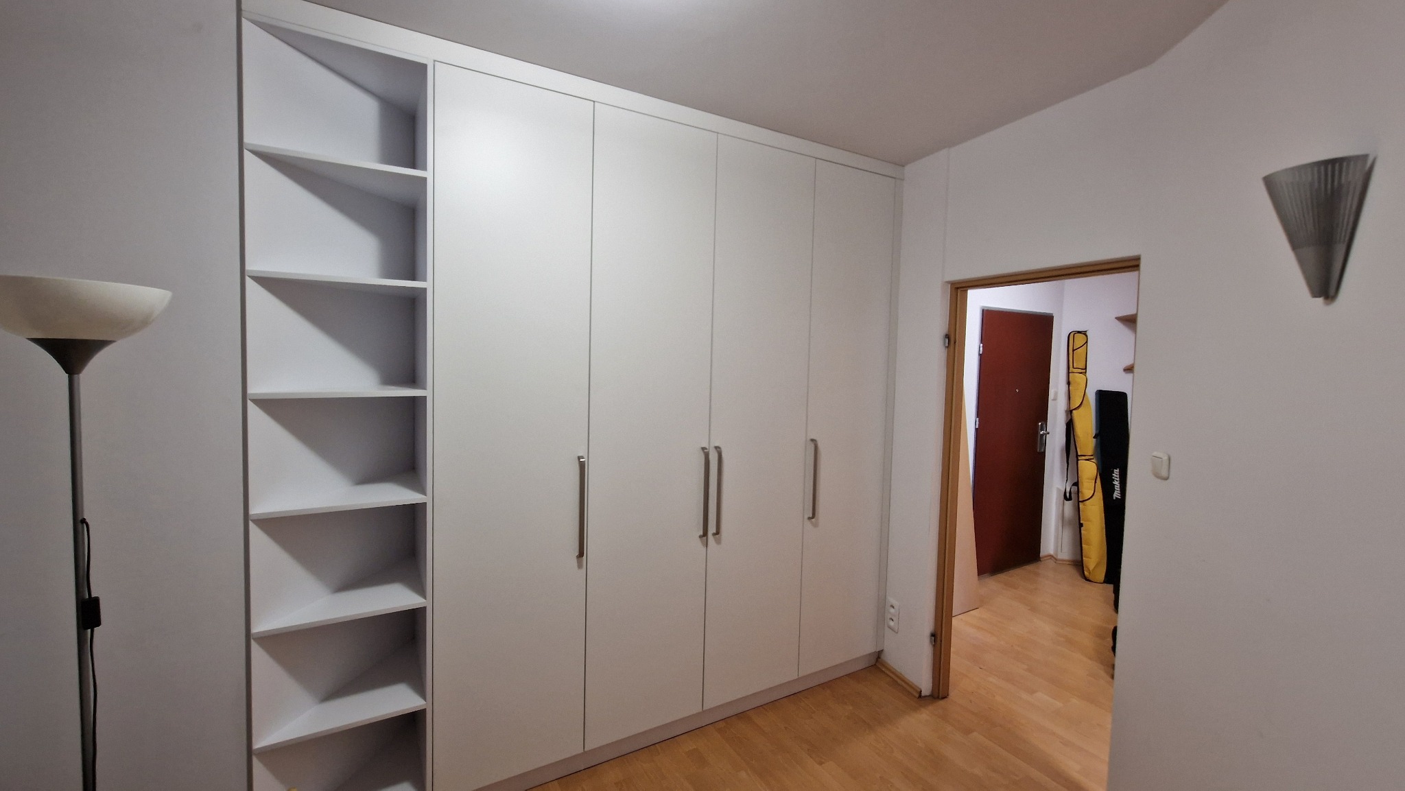 Biała szafa wnękowa z półkami, minimalistyczny design, długie uchwyty, widok z perspektywy wnętrza, lampa podłogowa w rogu, parkiet na podłodze.