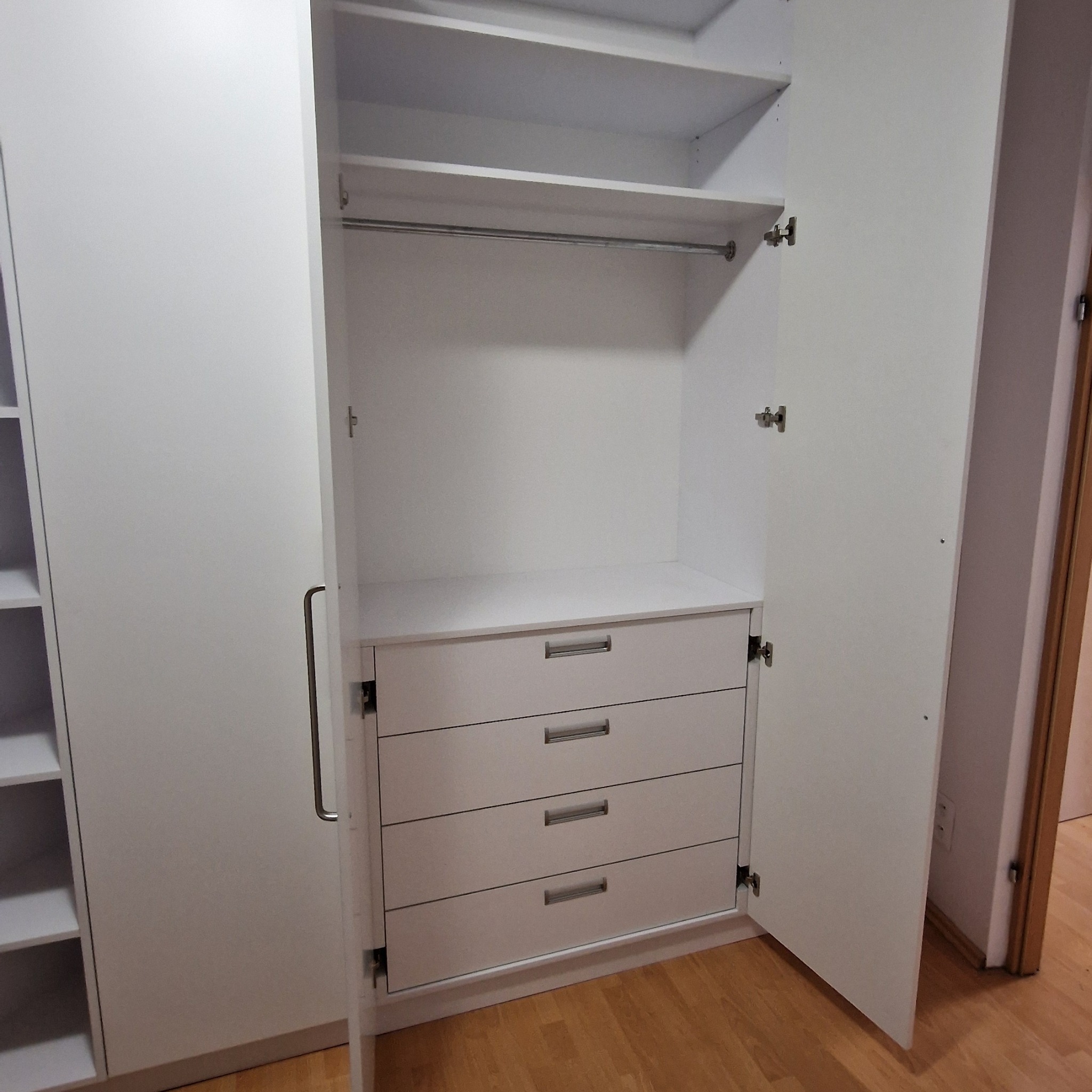 Biała szafa z czterema szufladami i drążkiem na ubrania, otwarte drzwi ukazują wnętrze. Minimalistyczny design, jasne oświetlenie, drewniana podłoga.