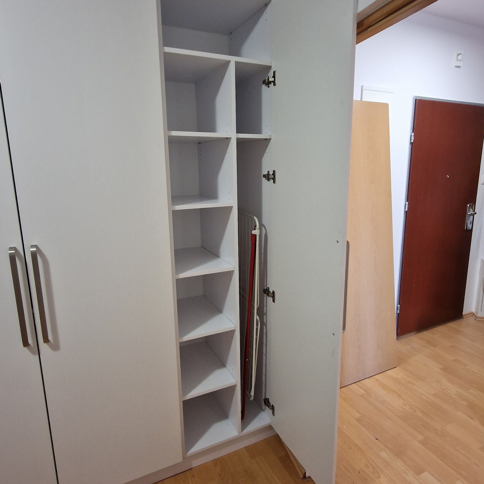 Biała szafa z otwartymi drzwiami, widoczne półki i deska do prasowania w środku. Minimalistyczny design, jasne wnętrze, drewniana podłoga.