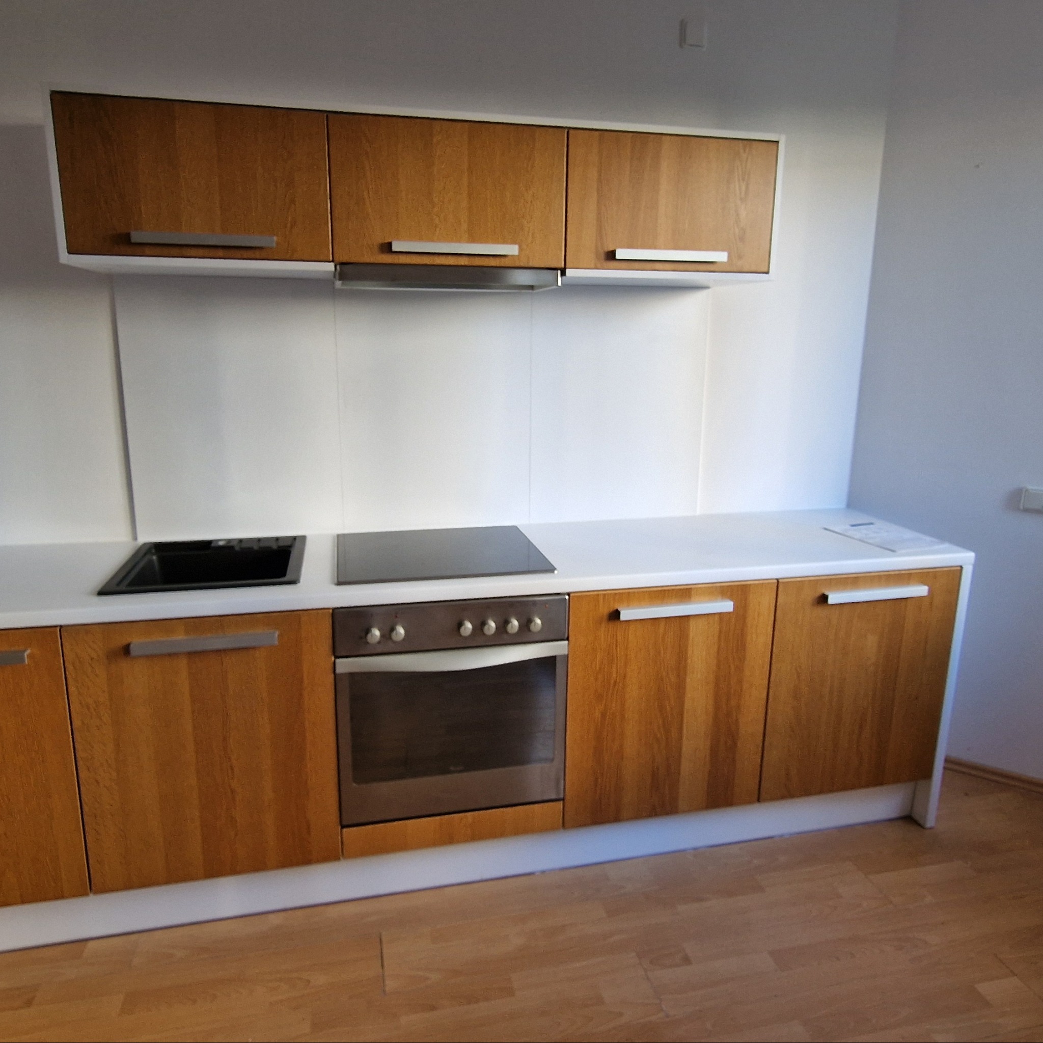 Jasna kuchnia z drewnianymi frontami szafek, białym blatem i sprzętem AGD. Minimalistyczny design, widok na dolne i górne szafki, nowoczesne uchwyty.