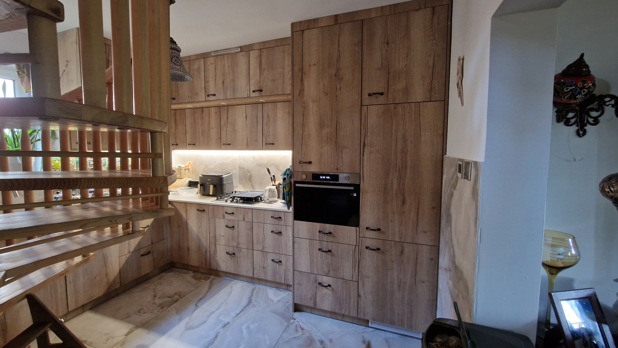 Jasna kuchnia z drewnianymi frontami i czarnymi uchwytami, widoczna z perspektywy drewnianych schodów. Na blacie kuchennym sprzęt AGD.