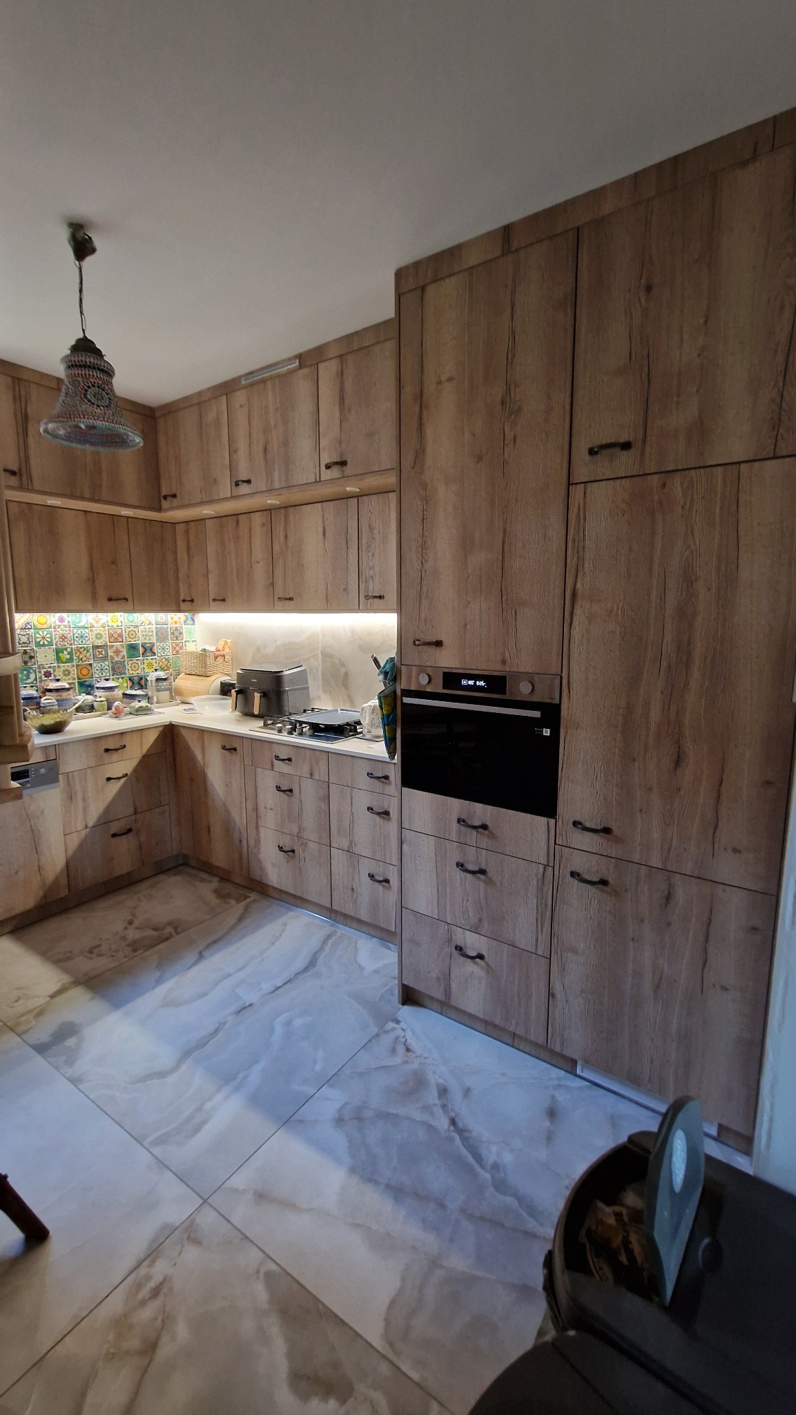 Jasna kuchnia z drewnianymi frontami, czarnymi uchwytami i marmurową podłogą. Uwagę przykuwa kolorowa mozaika nad blatem i designerska lampa.