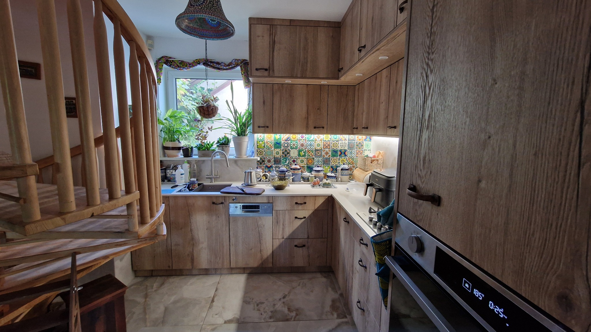 Kuchnia na wymiar z drewnianymi frontami, kolorową mozaiką nad blatem i spiralnymi schodami w tle. Ujęcie z perspektywy, widoczne wyposażenie i dekoracje.