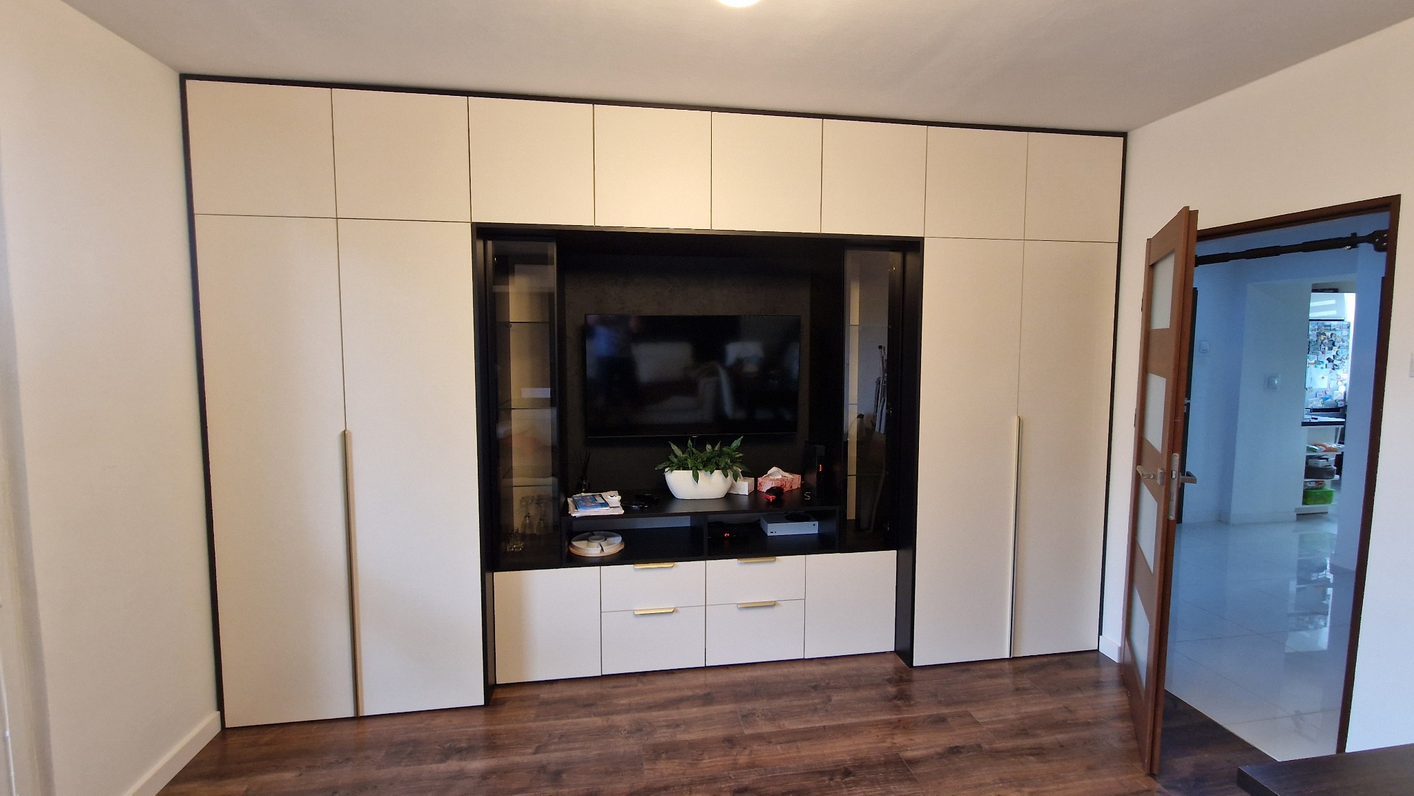 Biała zabudowa meblowa z szafkami i półkami, wnęka na telewizor, czarne obramowanie, drewniana podłoga, otwarte drzwi do innego pomieszczenia.