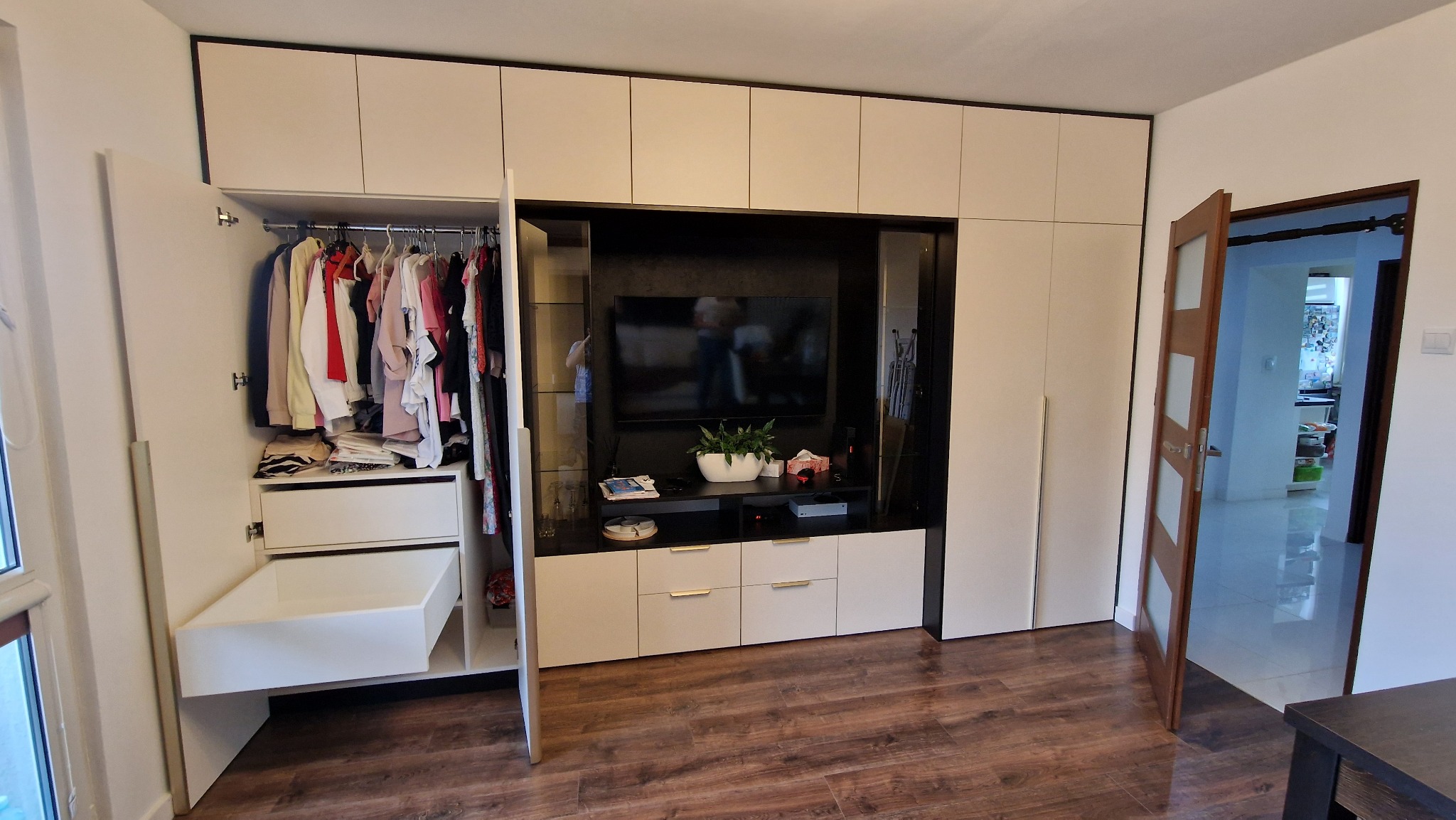 Biała zabudowa meblowa z szafą z otwartymi drzwiami, telewizorem i szafkami RTV na tle parkietu. Widok na otwarte drzwi do innego pomieszczenia.