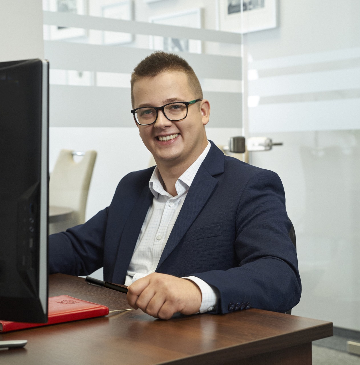 Uśmiechnięty doradca w okularach i granatowej marynarce siedzi za biurkiem z czerwonym notatnikiem i długopisem w biurze w Osielsku, widoczny monitor komputera.