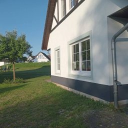 Tomasz Derwisz MTM ITA Krakow - Biały dom z szarymi akcentami i brązowym dachem, widok z trawnika. Okna z białymi ramami, rynna. W tle inne domy i zieleń. Słoneczny dzień.