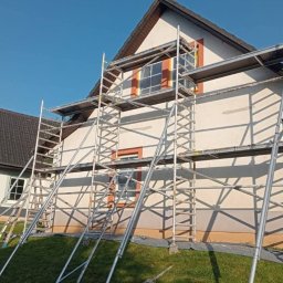 Tomasz Derwisz MTM ITA Krakow - Dom jednorodzinny w trakcie prac elewacyjnych, otoczony rusztowaniem. Widoczne fragmenty ocieplenia i świeżo pomalowana fasada. Słoneczny dzień.