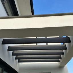 Tomasz Derwisz MTM ITA Krakow - Nowoczesna pergola z czarnymi belkami na tle jasnej elewacji i błękitnego nieba. Widok z dołu, podkreślający minimalistyczny design i precyzję wykonania.