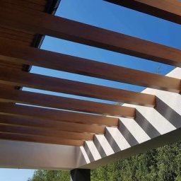 Tomasz Derwisz MTM ITA Krakow - Nowoczesna pergola z drewnianymi belkami i białym wykończeniem, widziana z dołu na tle błękitnego nieba i zieleni drzew. Minimalistyczny design i gra światła.