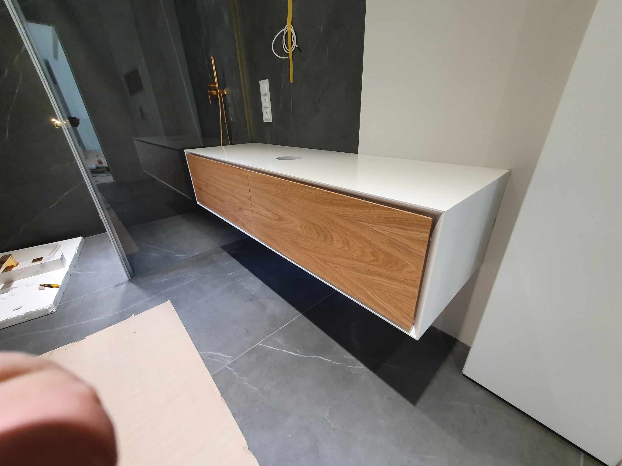 Nowoczesna szafka łazienkowa pod umywalkę z drewnianymi frontami i białym blatem, zamontowana na ścianie z szarymi płytkami imitującymi marmur, widoczne przewody elektryczne i elementy instalacji...