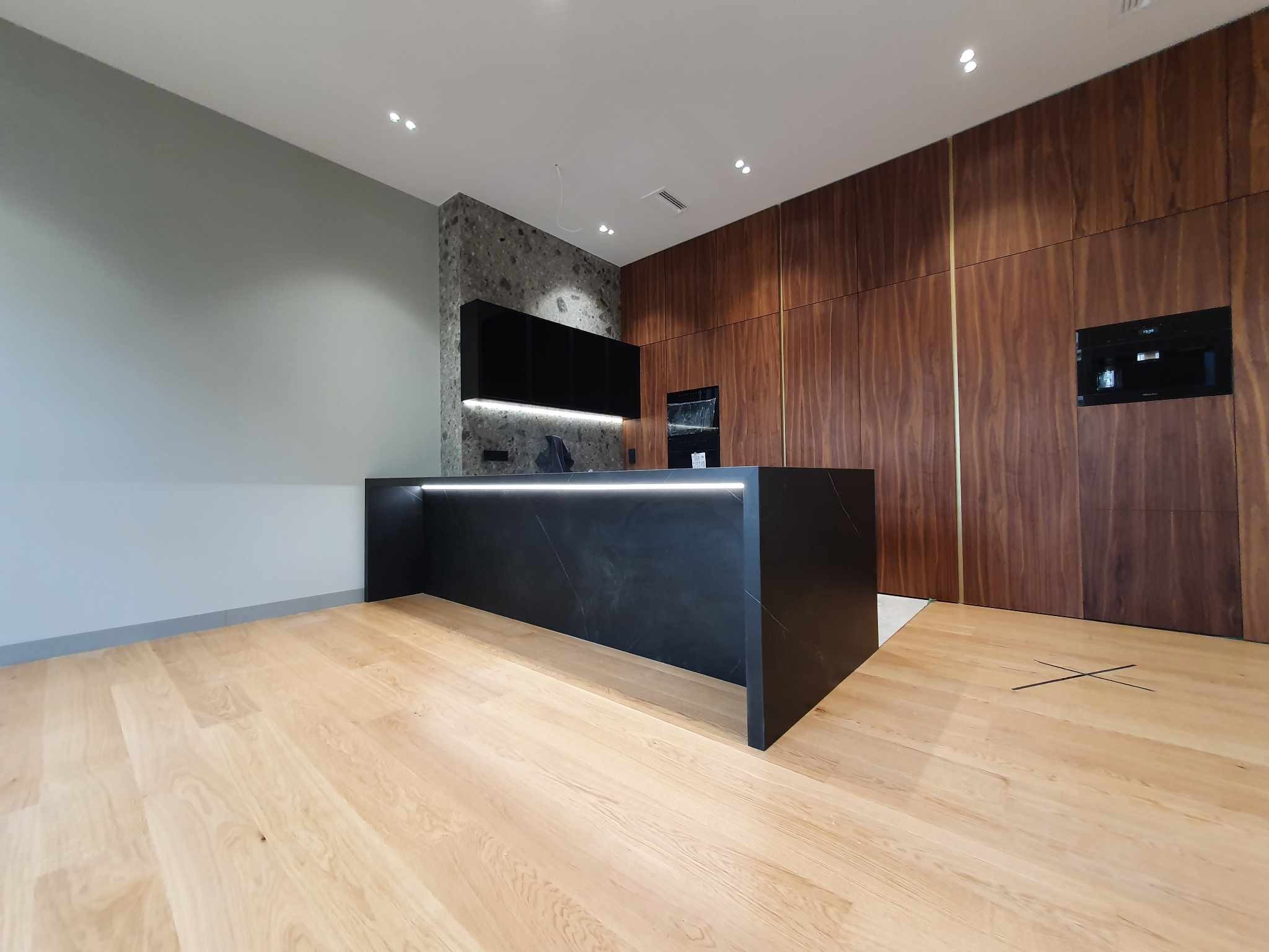 Nowoczesna kuchnia z czarnym blatem wyspowym z podświetleniem LED, drewnianą zabudową z piekarnikiem i kamienną ścianą z czarnymi szafkami wiszącymi.