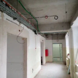 Wnętrze w trakcie remontu: widoczne koryta kablowe pod sufitem, przewody elektryczne w kolorach, ściany w stanie surowym, okno z materiałami budowlanymi na parapecie. Czerwona skrzynka na ścianie...