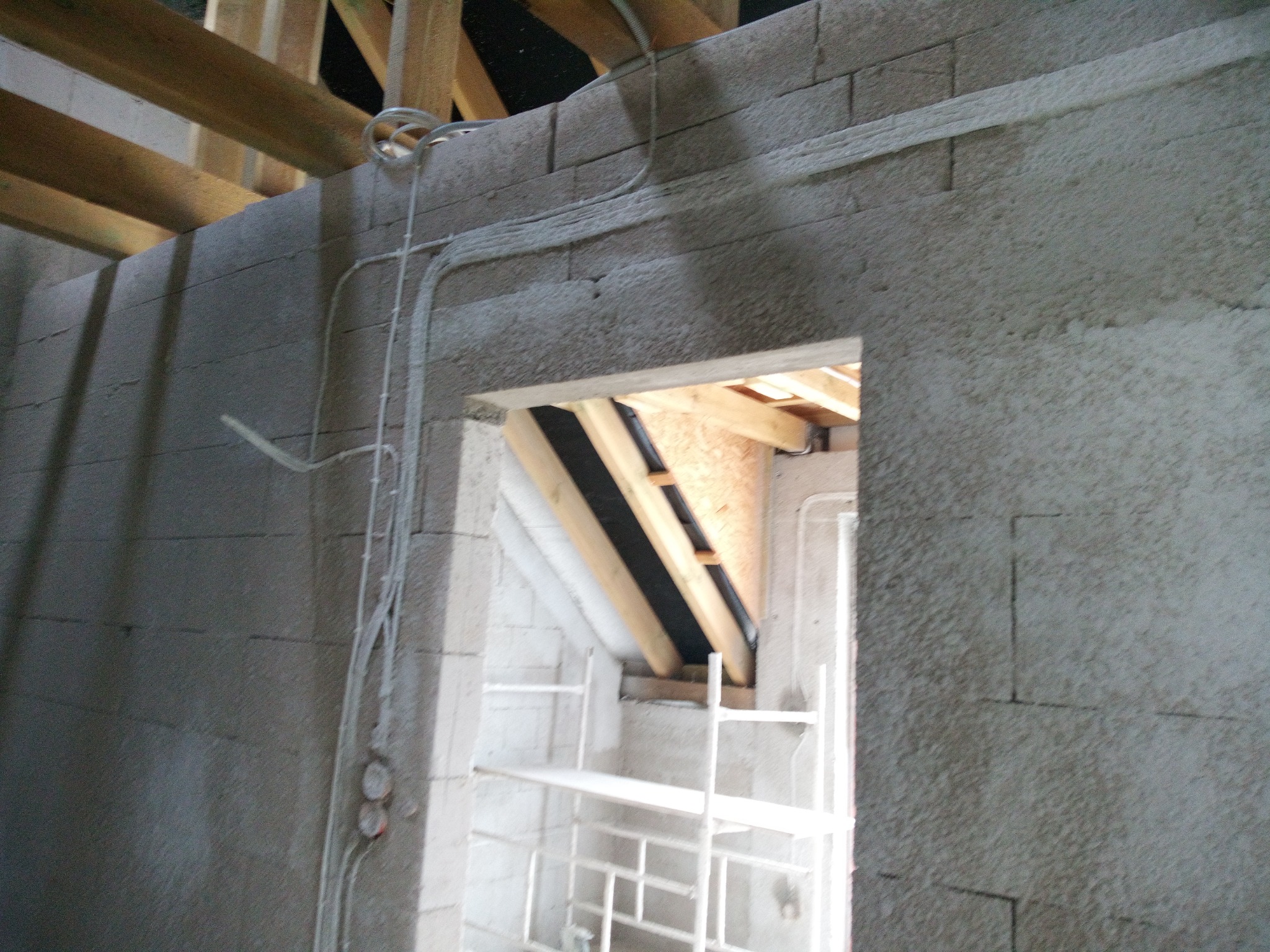 Surowe wnętrze w trakcie budowy z widocznymi przewodami elektrycznymi poprowadzonymi po ścianie z betonowych bloczków, drewnianą konstrukcją dachu i rusztowaniem w tle.