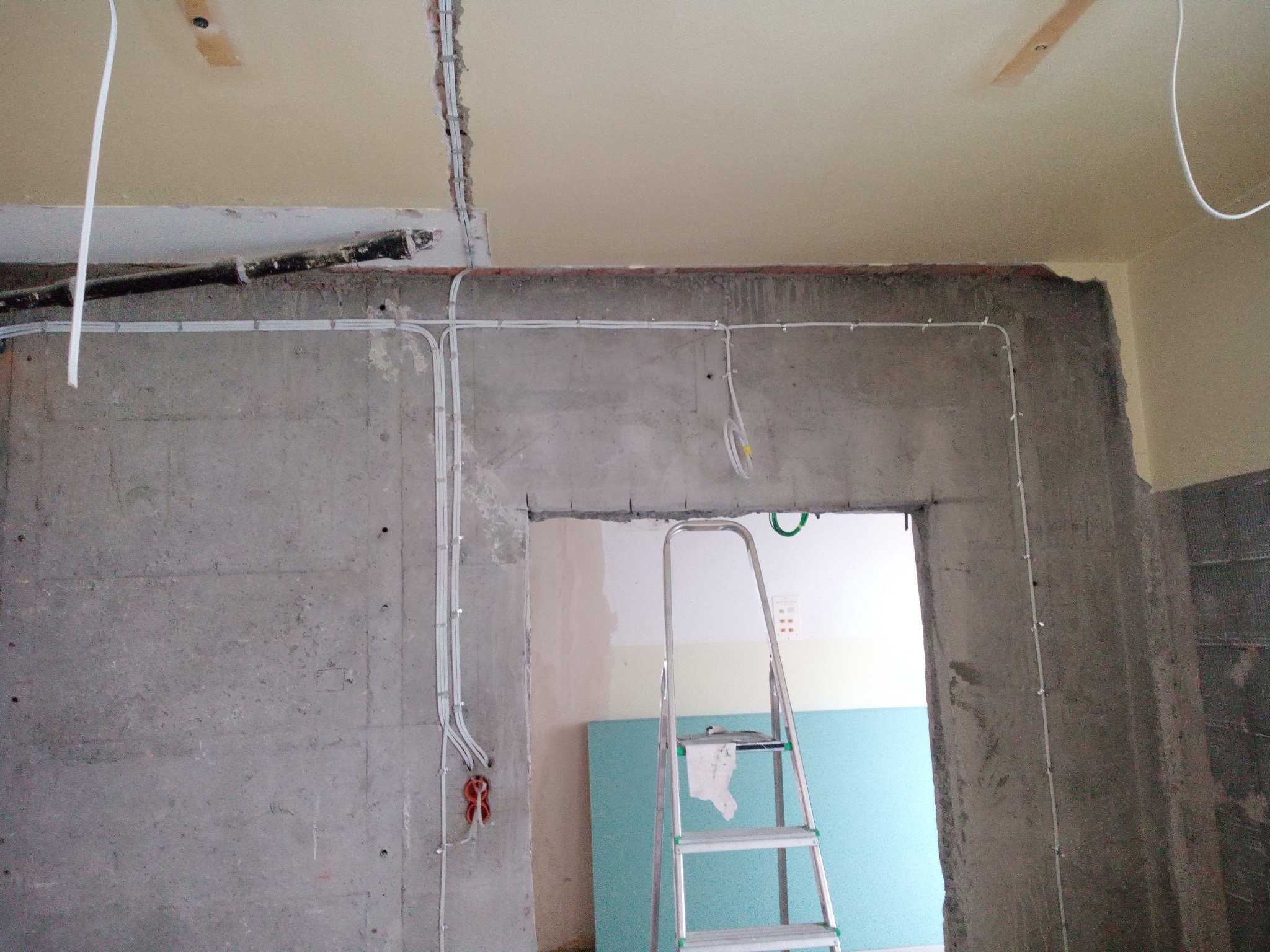 Surowy betonowy pokój w trakcie remontu z widocznymi przewodami elektrycznymi prowadzonymi po ścianach, drabina aluminiowa oparta o ścianę, w tle niebieska płyta izolacyjna.