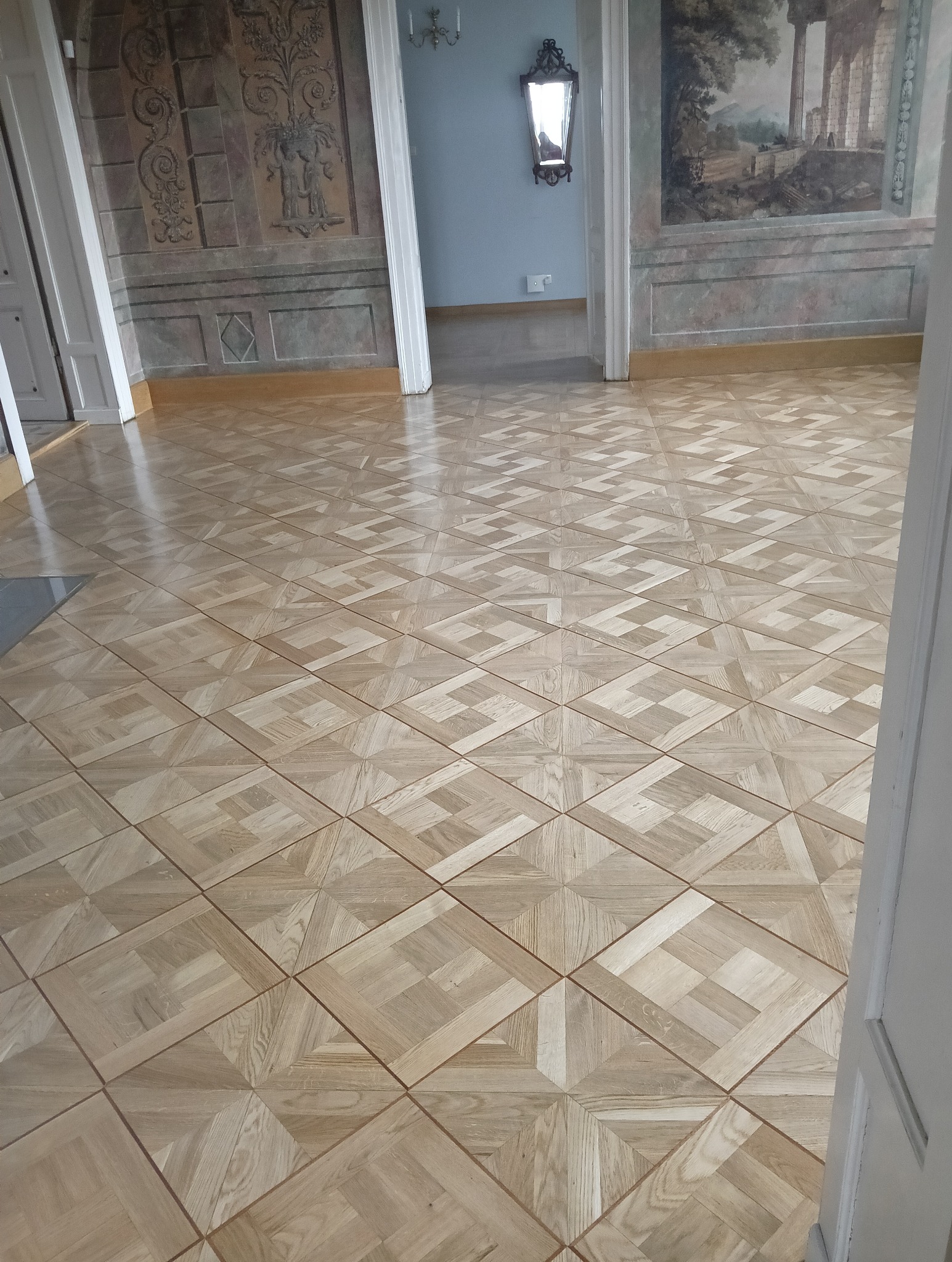 Jasny parkiet ułożony we wzór kwadratów i rombów w przestronnym wnętrzu z dekoracyjnymi malowidłami ściennymi i lustrem.