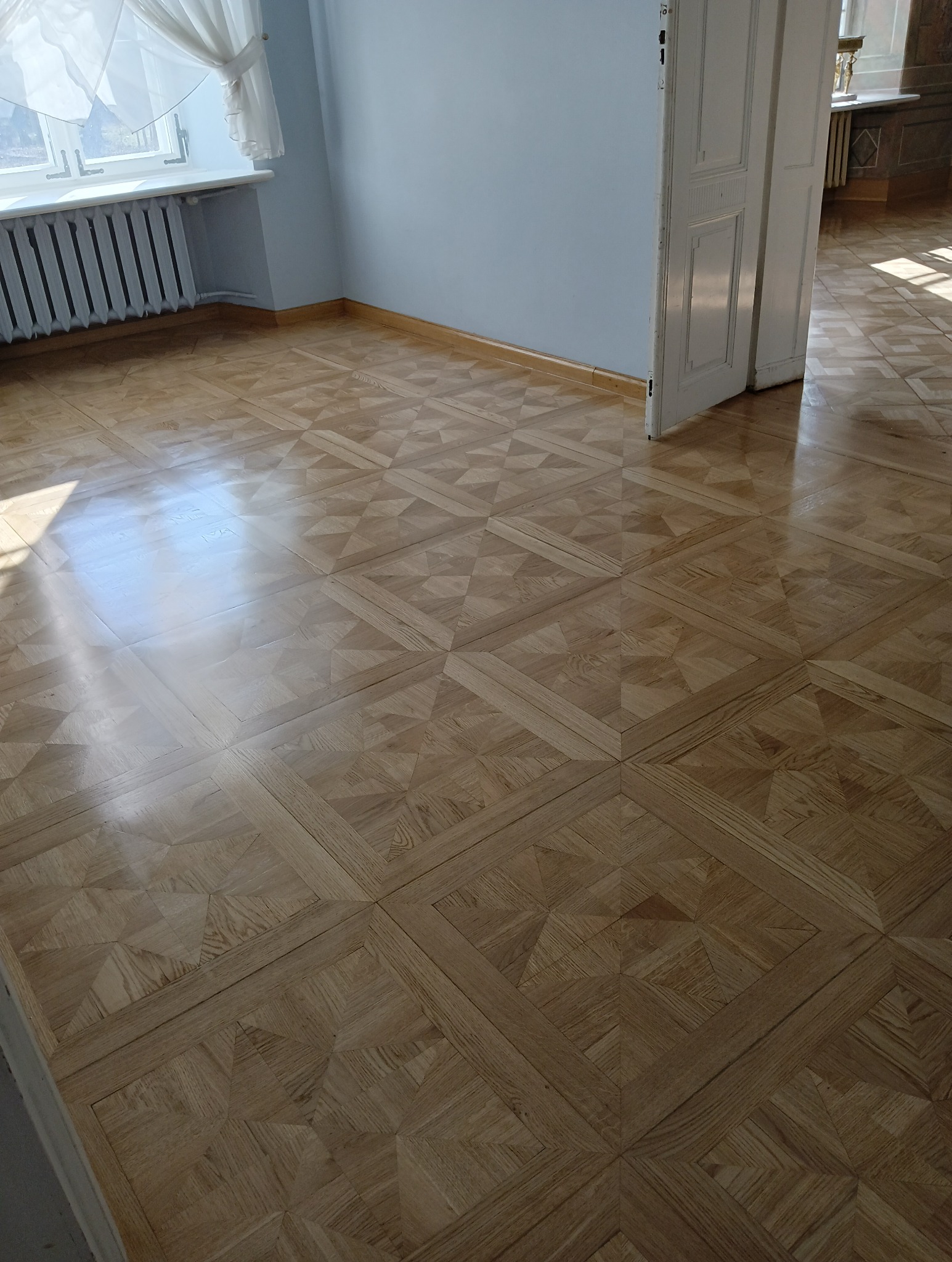 Parkiet ułożony we wzór kwadratów z wypełnieniem w jodełkę, widok na jasne, wycyklinowane drewno w pokoju z oknem i grzejnikiem.