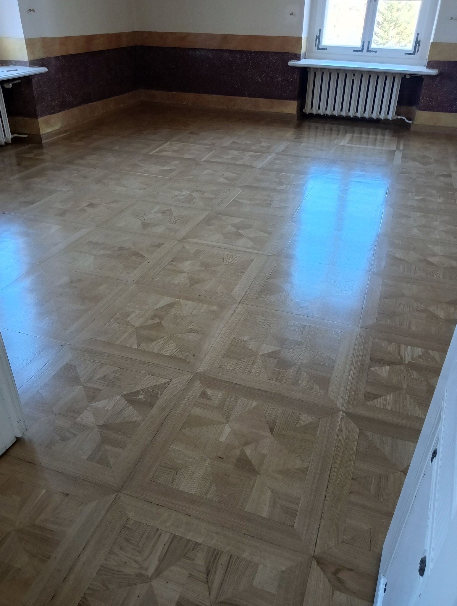 Jasny parkiet ułożony we wzór kwadratów z wewnętrznymi trójkątami, widoczny fragment ściany z bordową lamperią i grzejnik pod oknem, odbicie światła na podłodze.