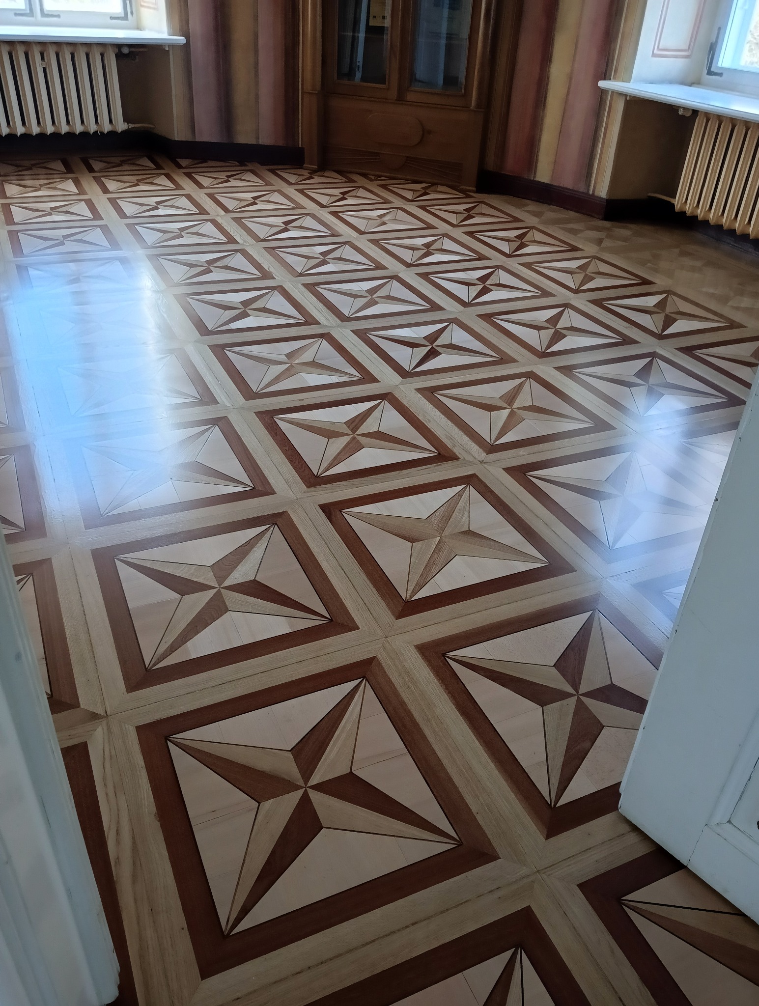 Wypolerowana podłoga z parkietu w geometryczny wzór gwiazdy, widoczne grzejniki i fragmenty drzwi.