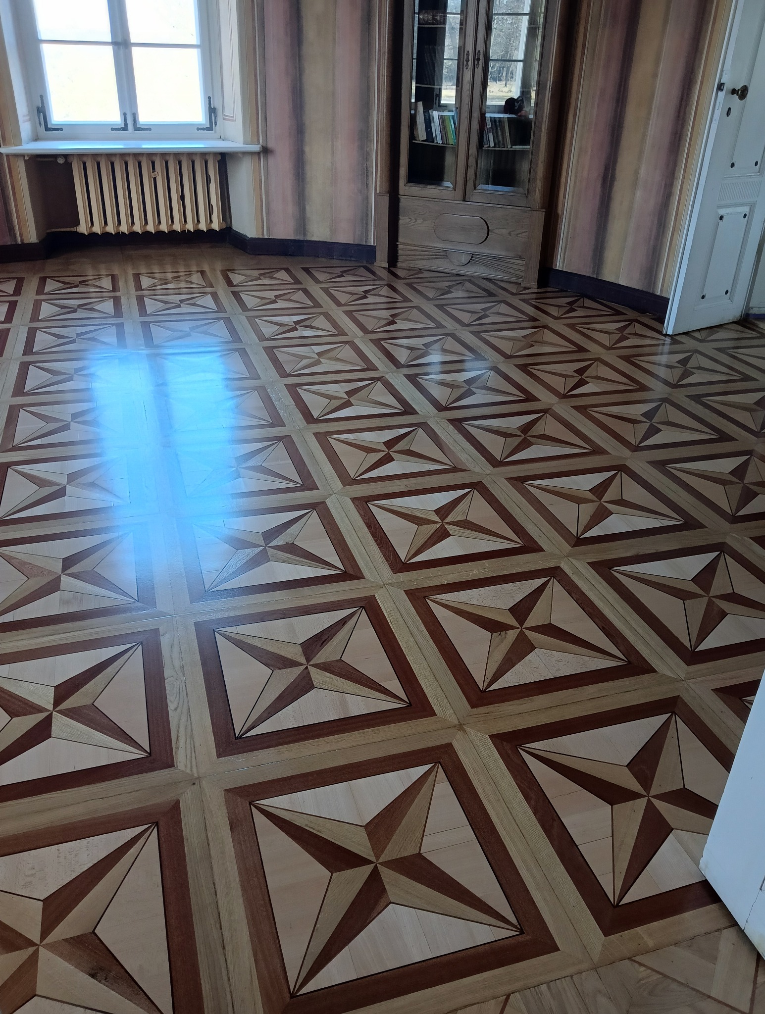 Parkiet z wzorem gwiazdy w kwadracie, ułożony w klasycznym wnętrzu z grzejnikiem i szafą w tle, widoczny odblask światła słonecznego na podłodze.