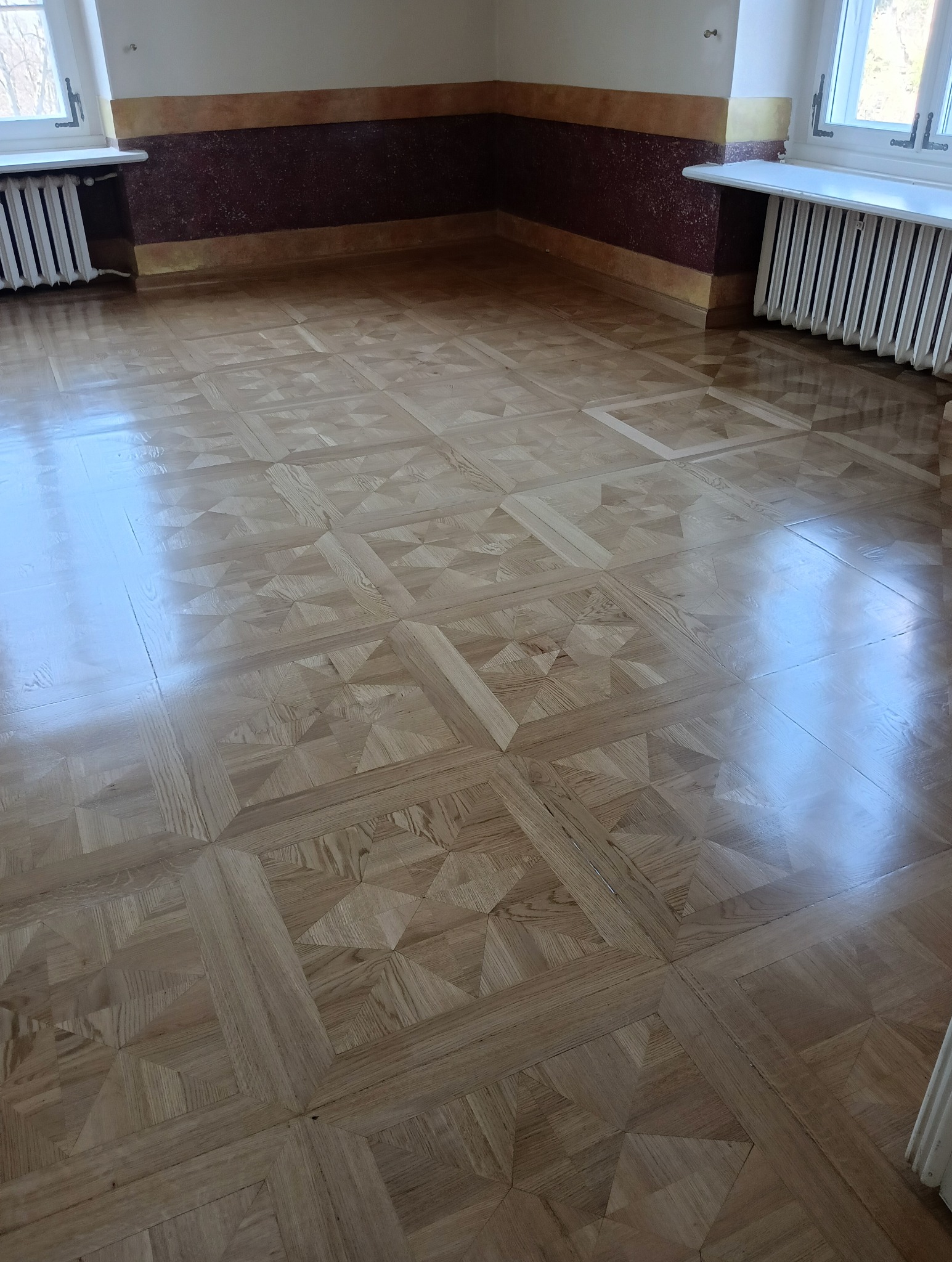 Błyszcząca, nowo położona podłoga parkietowa z geometrycznym wzorem, widok na fragment pokoju z grzejnikami i oknami.