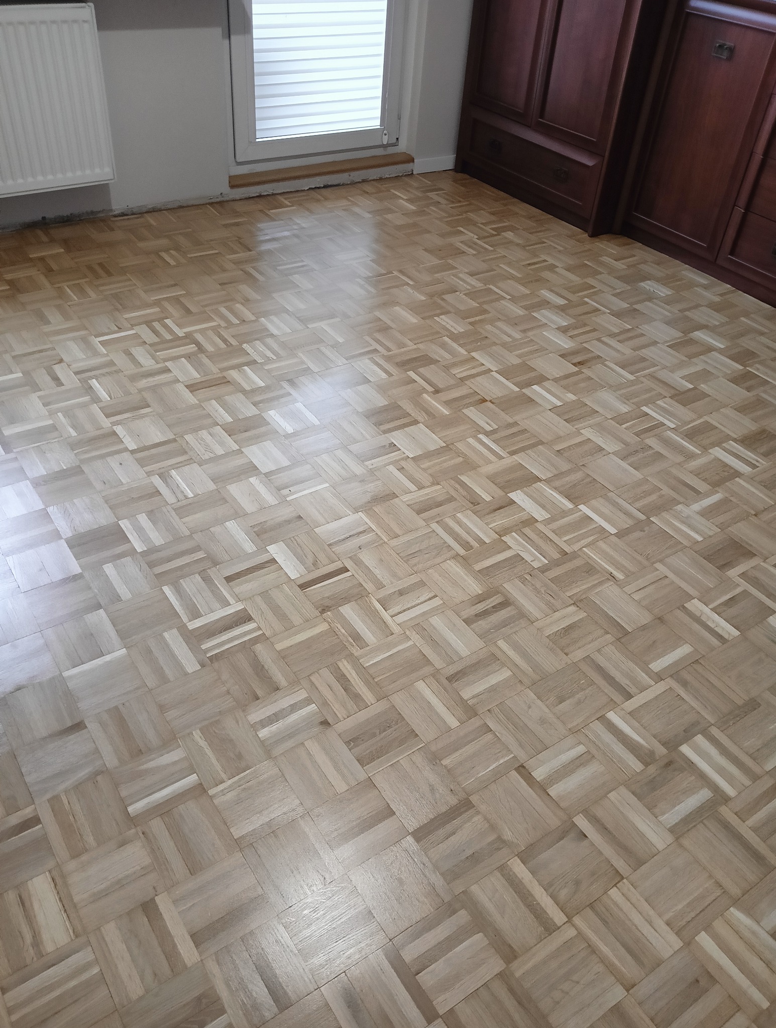 Odnowiona podłoga z parkietu w jasnym odcieniu drewna, ułożona we wzór kwadratów, widoczne okno z żaluzjami i ciemne drewniane meble w tle.