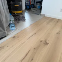 Jasne panele podłogowe z widoczną strukturą drewna, fragment odkurzacza przemysłowego i butelka wody w tle, po zakończeniu montażu.