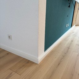 GoodWood Wilhelmus Aarts - Jasne panele podłogowe z listwami przypodłogowymi, stykające się z białą i zieloną ścianą o wyraźnej fakturze, gniazdko elektryczne przy podłodze.