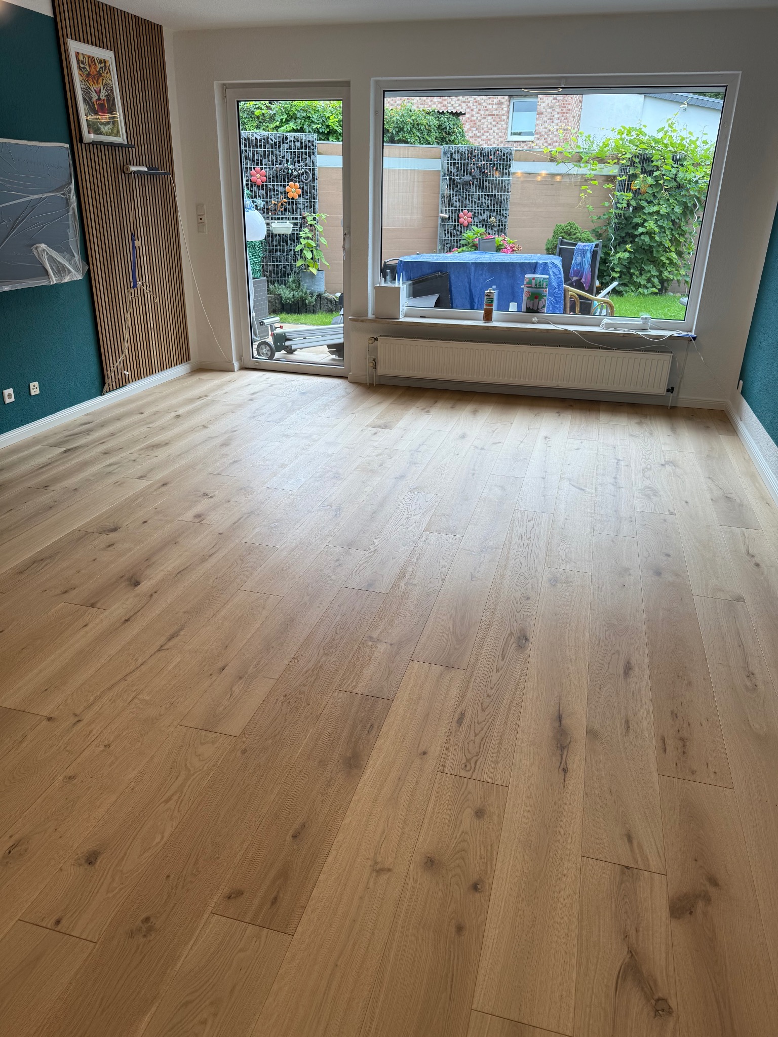 Jasna, drewniana podłoga w pokoju z dużym oknem na ogród. Ściana w kolorze morskim z drewnianymi panelami. Widoczny fragment okna balkonowego.