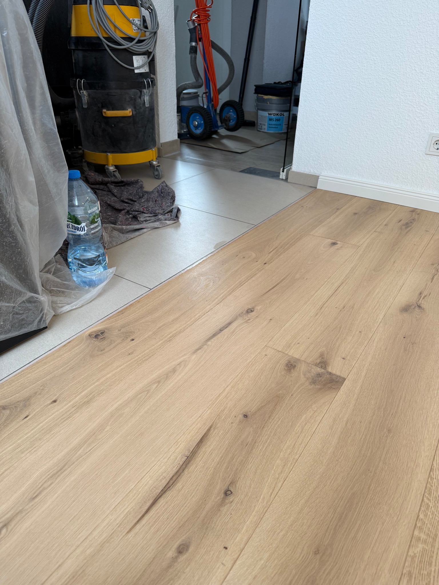 Jasne panele podłogowe z widoczną strukturą drewna, fragment odkurzacza przemysłowego i butelka wody w tle, po zakończeniu montażu.